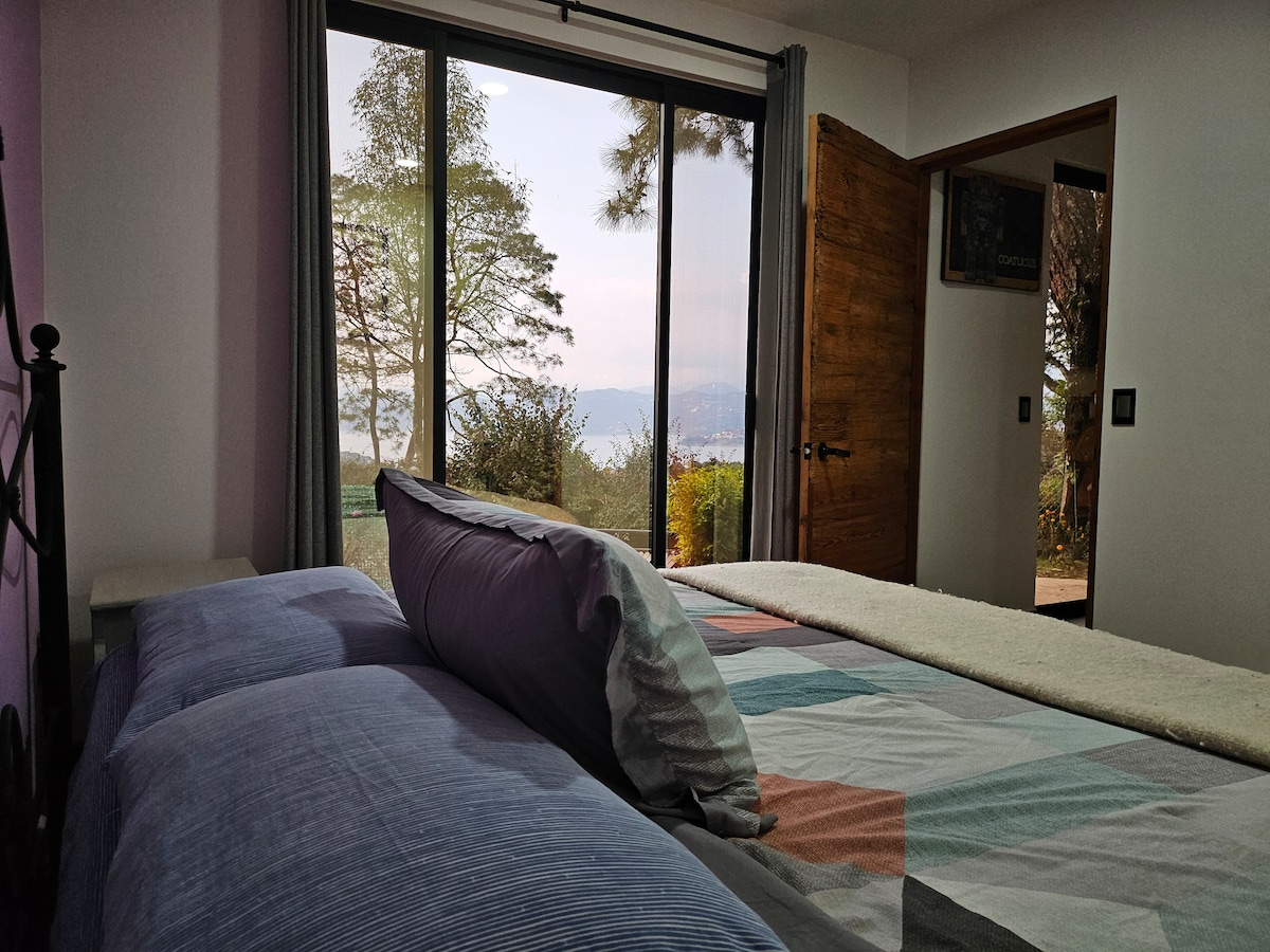 A spacious bedroom features a large window offering views of the surrounding landscape. A cozy bed is adorned with a patterned blanket and multiple pillows. The room includes a nightstand beside the bed, and a wooden door leading to another area is visible.
