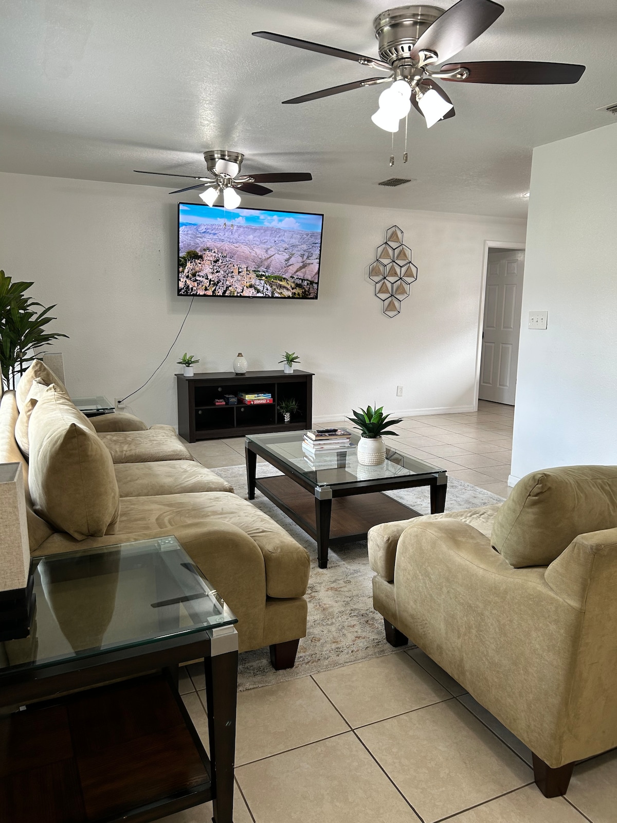 A bright living area features a comfortable beige sofa set and a dark coffee table. A wall-mounted television displays scenic imagery above a modern entertainment unit. Indoor plants add a touch of greenery, and ceiling fans enhance airflow in the space.