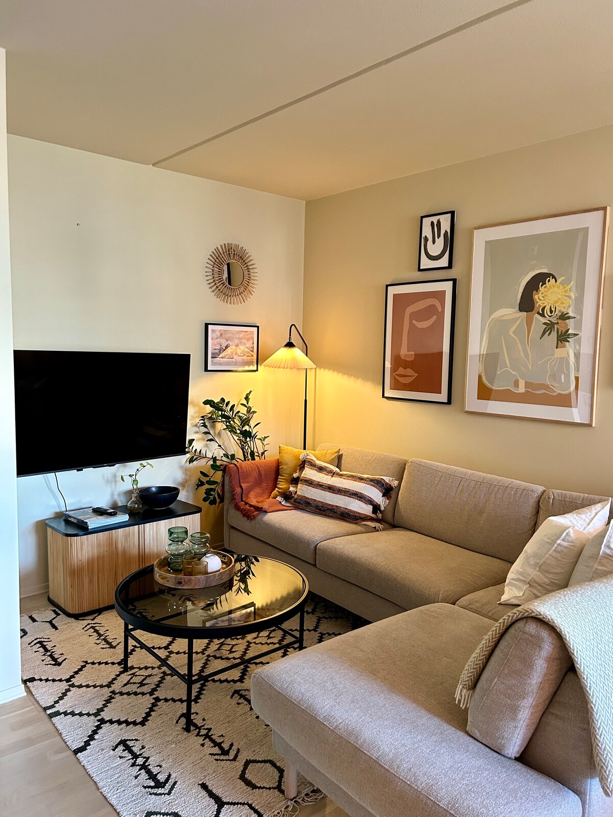 A modern living area features a comfortable sectional sofa adorned with decorative pillows. A round coffee table sits in front, complemented by a textured rug below. The wall is decorated with framed artwork, and a flat-screen TV is mounted on the opposite wall, adding to the room's inviting atmosphere.