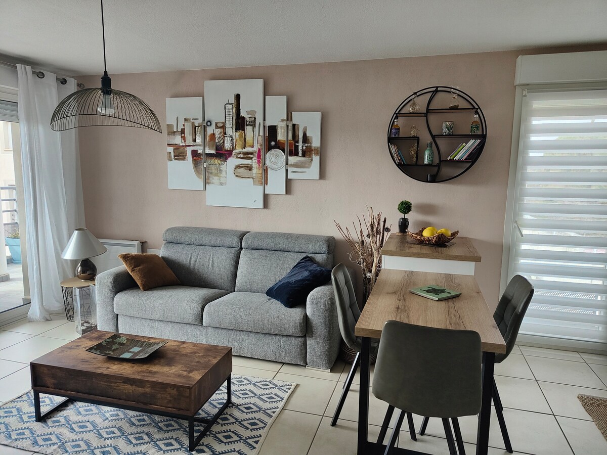 A modern living area is shown, featuring a comfortable gray sofa and a wooden coffee table. A rectangular dining table with chairs is positioned nearby. Wall art depicting cityscape elements is displayed above the sofa. Natural light filters through sheer window coverings.