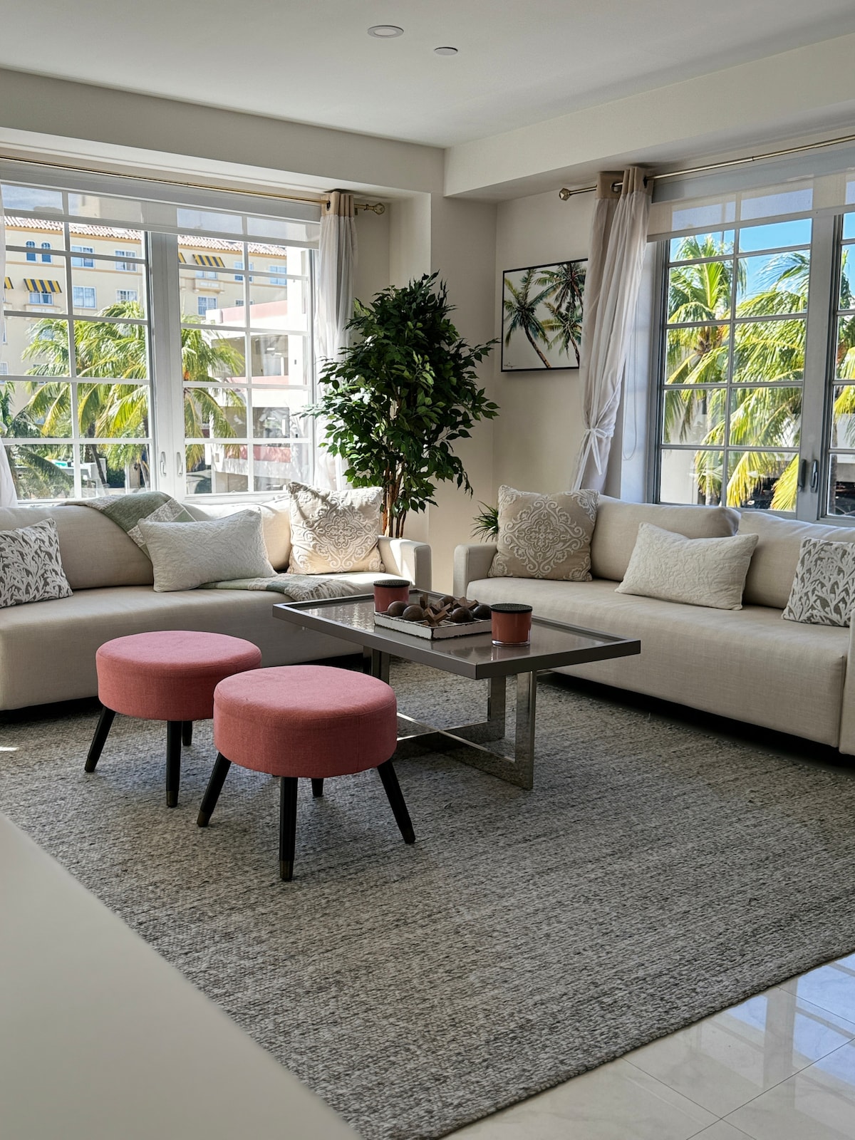 A light-filled living area is furnished with two sofas adorned with decorative pillows and a low coffee table. A pair of pink stools adds a pop of color, while large windows frame views of swaying palm trees outside. Soft area rugs enhance the cozy atmosphere.