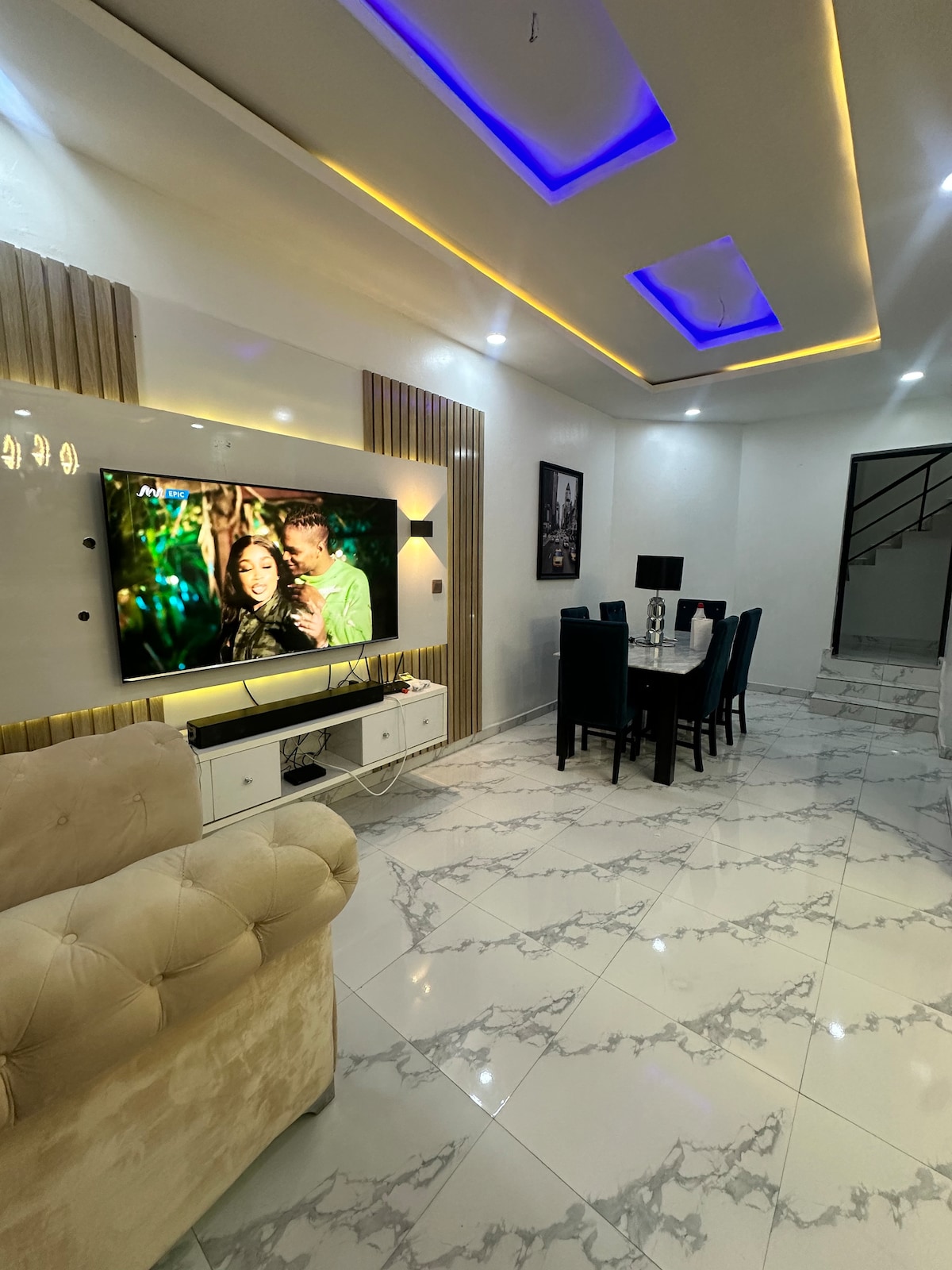 A modern living area features a large television mounted on a sleek wall unit. Elegant dining furniture, including a table with six chairs, is arranged nearby. The space is illuminated by recessed lighting and has a polished marble floor adding to the contemporary design.