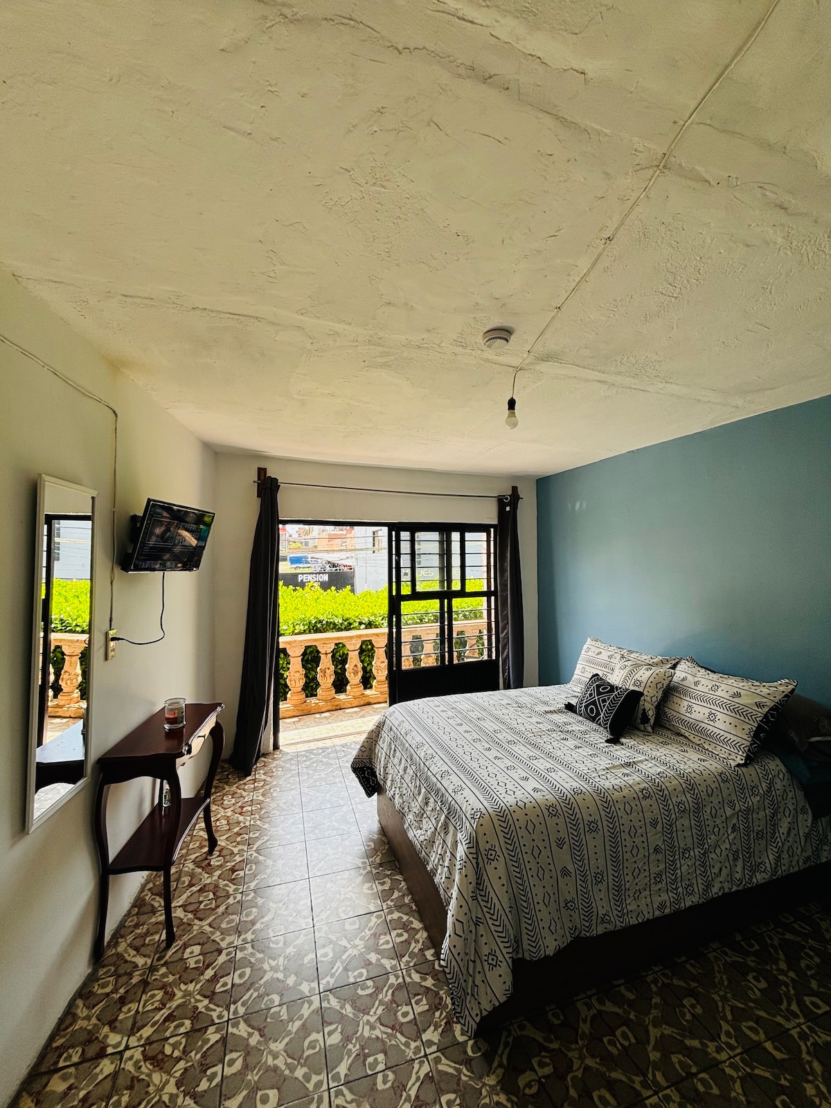A comfortable bedroom features a spacious bed with decorative bedding. A large window allows natural light to illuminate the room and offers views of the balcony. A wall-mounted TV is visible, alongside a small table and mirror, enhancing the room's functionality.
