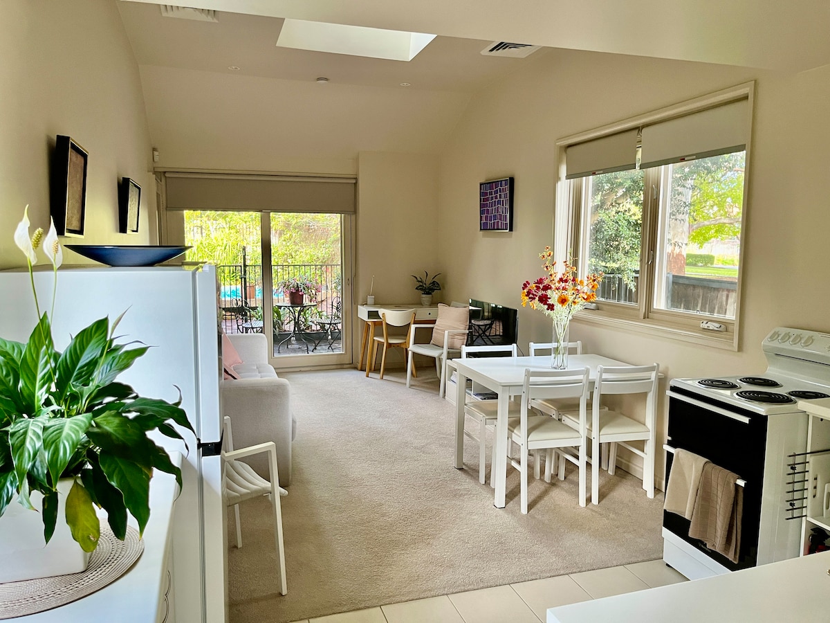 A bright living area features a light-colored carpet and ample natural light from large windows. A dining table with chairs is positioned next to a seating area, while a full kitchen with modern appliances is visible. Lush greenery can be seen through the glass doors leading to a private outdoor space.