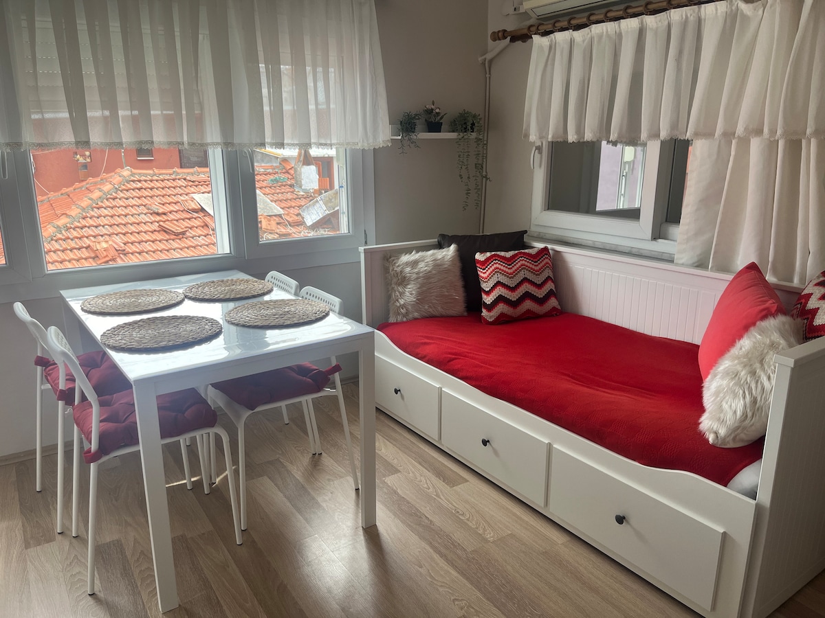 A bright living area features a white dining table surrounded by four chairs, complemented by woven placemats. A cozy daybed with a red blanket and patterned pillows sits nearby, with large windows dressed in sheer curtains allowing natural light to fill the space.
