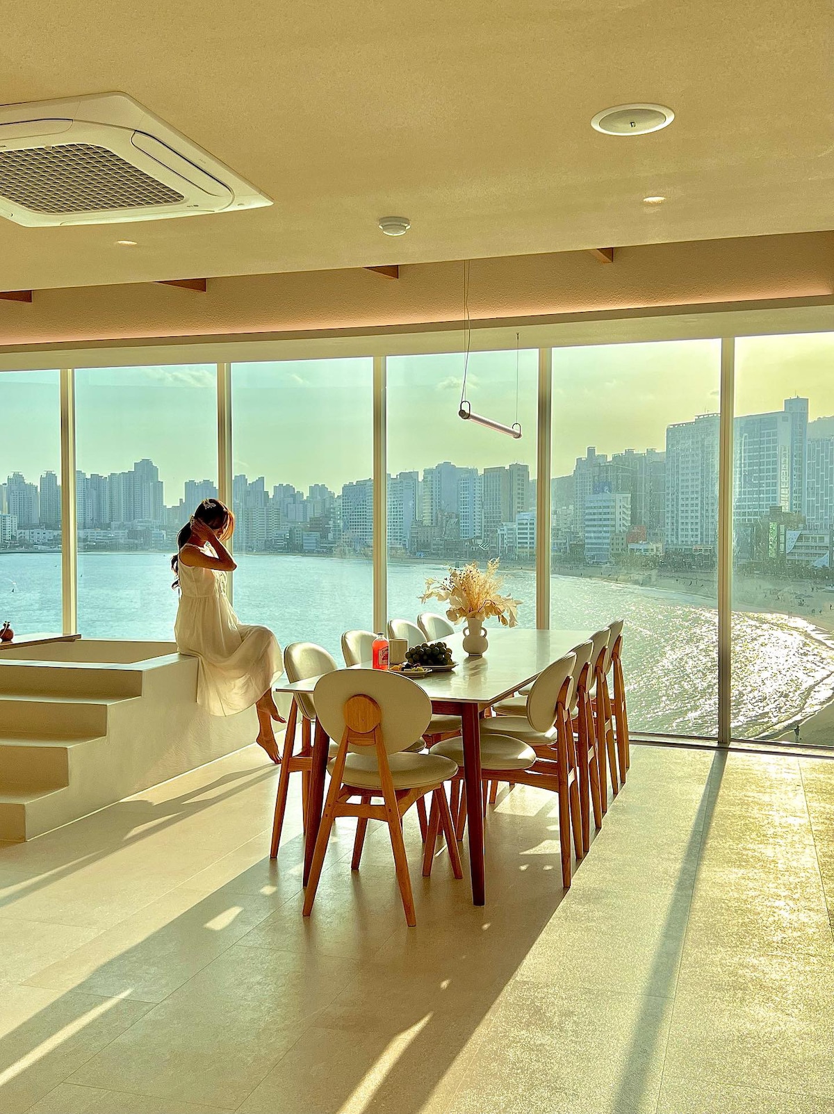 A spacious dining area is complemented by a large window offering a view of the city skyline and the beach. A modern table is surrounded by stylish chairs, while soft light reflects off the water, creating a serene atmosphere.