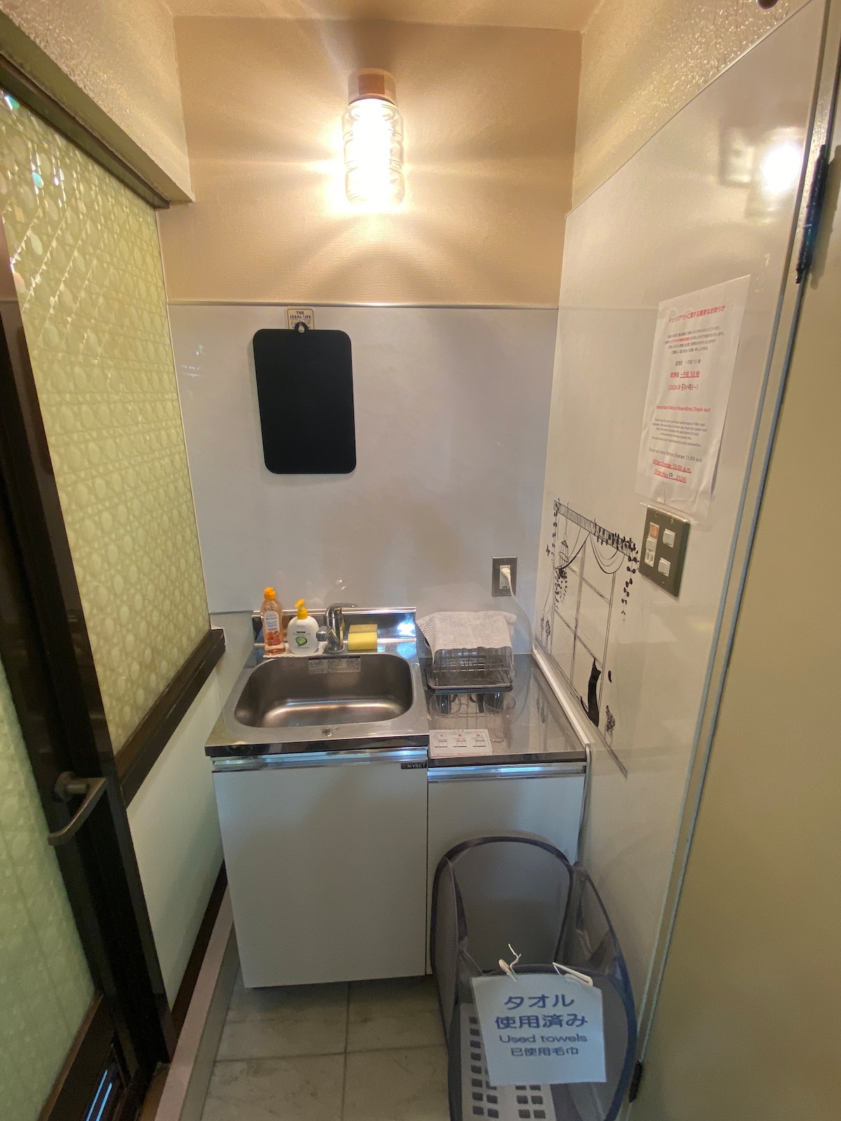 The compact kitchenette is equipped with a small sink and cooking area. Condiments and dish soap are visible on the countertop. A cabinet below provides additional storage. A laminated sign about towel usage is displayed on the wall, and a trash bin is positioned nearby.