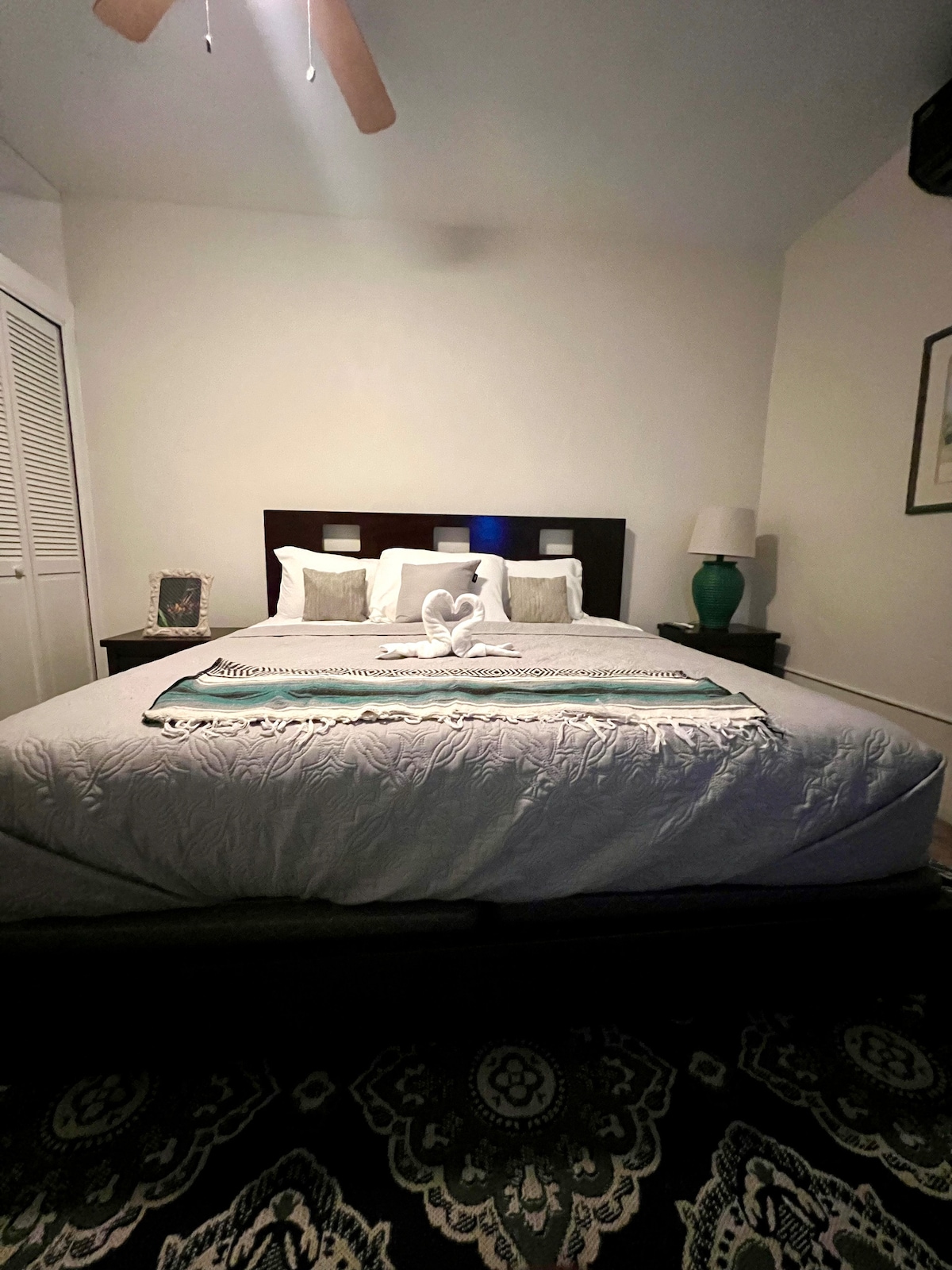 A spacious king-sized bed is positioned in the center of the room, featuring soft white linens and a decorative throw blanket. Two bedside tables provide lighting, while a fan hangs from the ceiling. A tasteful rug accents the floor, and artwork adorns the walls.
