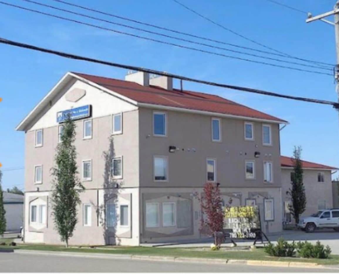 A three-story building features a light-colored exterior with large windows. Surrounding trees provide a sense of greenery, and a prominent sign is visible in front. The buildingās design is simple, with a red roof contrasting the neutral tones.