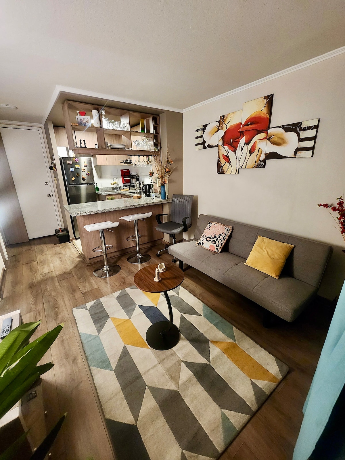 A welcoming living area features a gray sofa and a small coffee table atop a geometric-patterned rug. Shelving displays decorative items and kitchen essentials. A wall-mounted artwork of flowers is present, while a bright window curtain adds a touch of color to the space.
