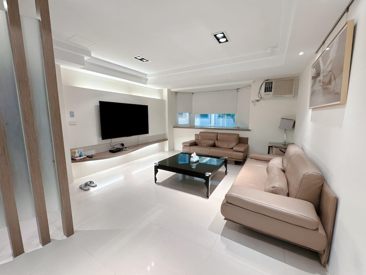 A modern living space features a flat-screen television mounted on the wall above a sleek, low coffee table. Comfortable leather sofas are arranged around the table, and soft lighting provides a warm ambiance. Large windows allow natural light to enter the bright, polished room.