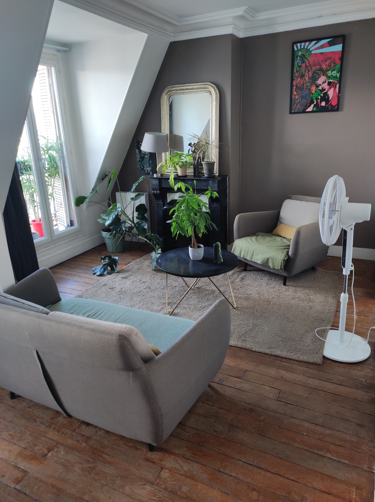 The living area features two comfortable armchairs, positioned around a circular coffee table. A decorative mirror adorns the wall, while potted plants bring a touch of greenery. Bright light streams through large windows, illuminating the wooden flooring and creating a cozy environment.