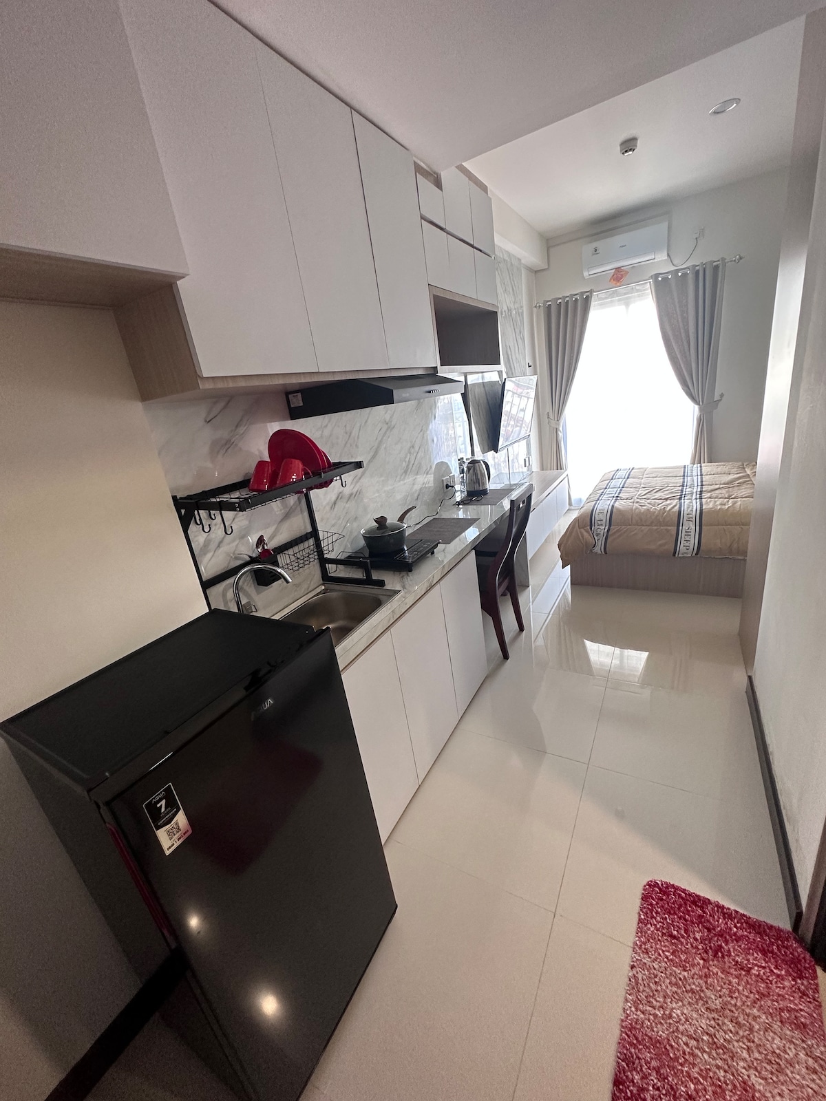 A compact kitchen area is equipped with modern appliances, including a refrigerator and cooking essentials. A cozy sleeping area with a neatly made bed is visible in the background. Natural light enters through a window, enhancing the airy feel of the space.