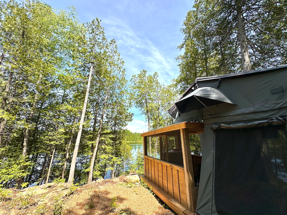 A spacious wooden platform extends from the side of a rooftop tent nestled among lush trees. A screened shelter with large windows offers views of the calm lake beyond, while an umbrella provides shade on the deck. The natural surroundings enhance the feeling of seclusion.