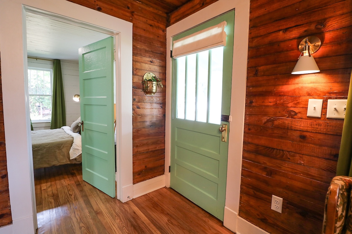 The entryway features original hardwood floors and shiplap walls, creating a rustic charm. Two green doors lead to the outside and an adjoining bedroom. A wall sconce provides soft lighting, enhancing the welcoming atmosphere. Natural light filters through windows adorned with simple curtains.