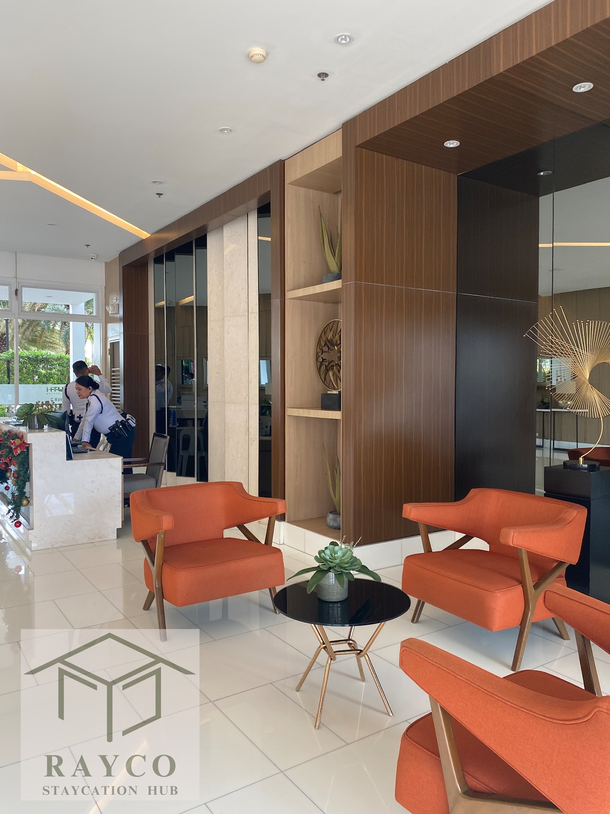 A modern lobby features stylish orange chairs paired with a round black table. Panoramic windows allow natural light to flow in, while decorative elements add elegance to the space. A front desk area is visible, providing a welcoming atmosphere for guests.