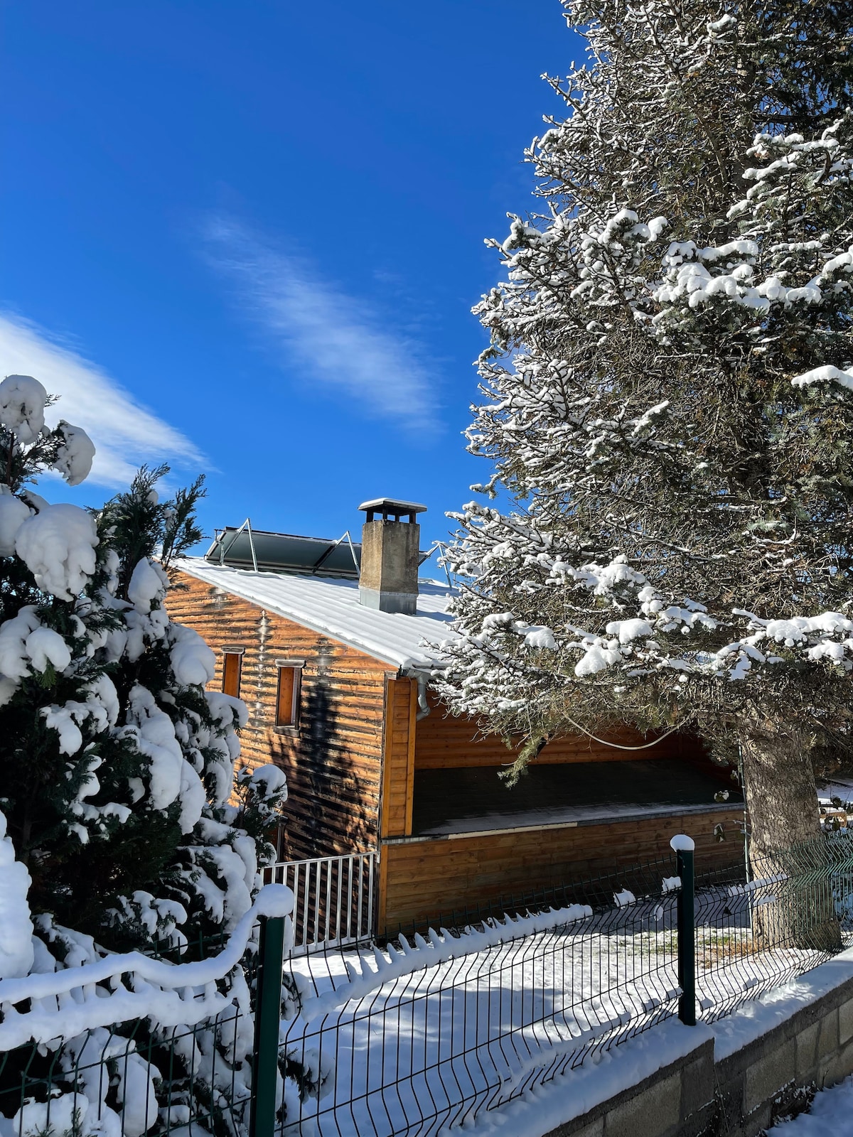 A wooden chalet is partially visible, surrounded by snow-covered trees. The building features a sloped roof with solar panels and a chimney. A protective fence outlines the property, while blue skies and fluffy clouds create a bright winter ambiance.