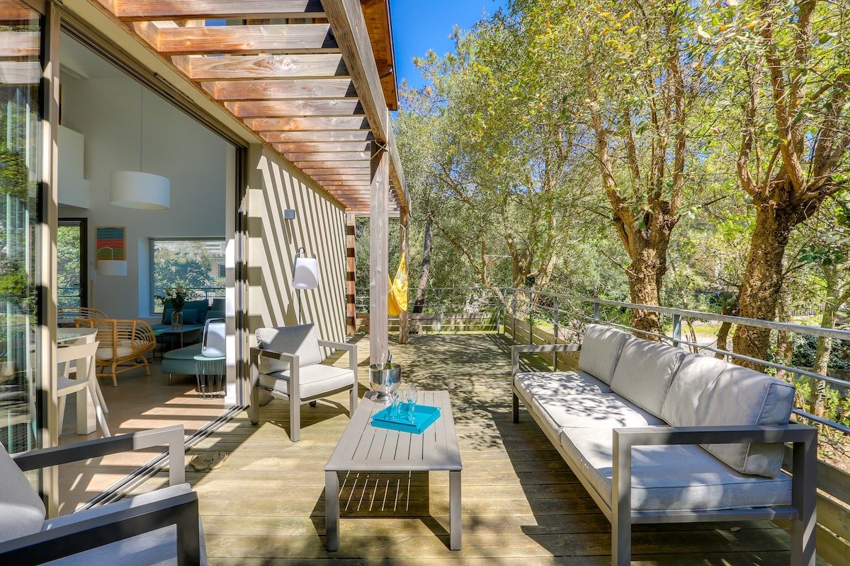An outdoor terrace features modern seating arrangements, including a gray sofa and armchairs. A wooden coffee table sits in the center. Surrounding trees provide shade and greenery, with large glass doors leading into the interior space, inviting natural light and views.