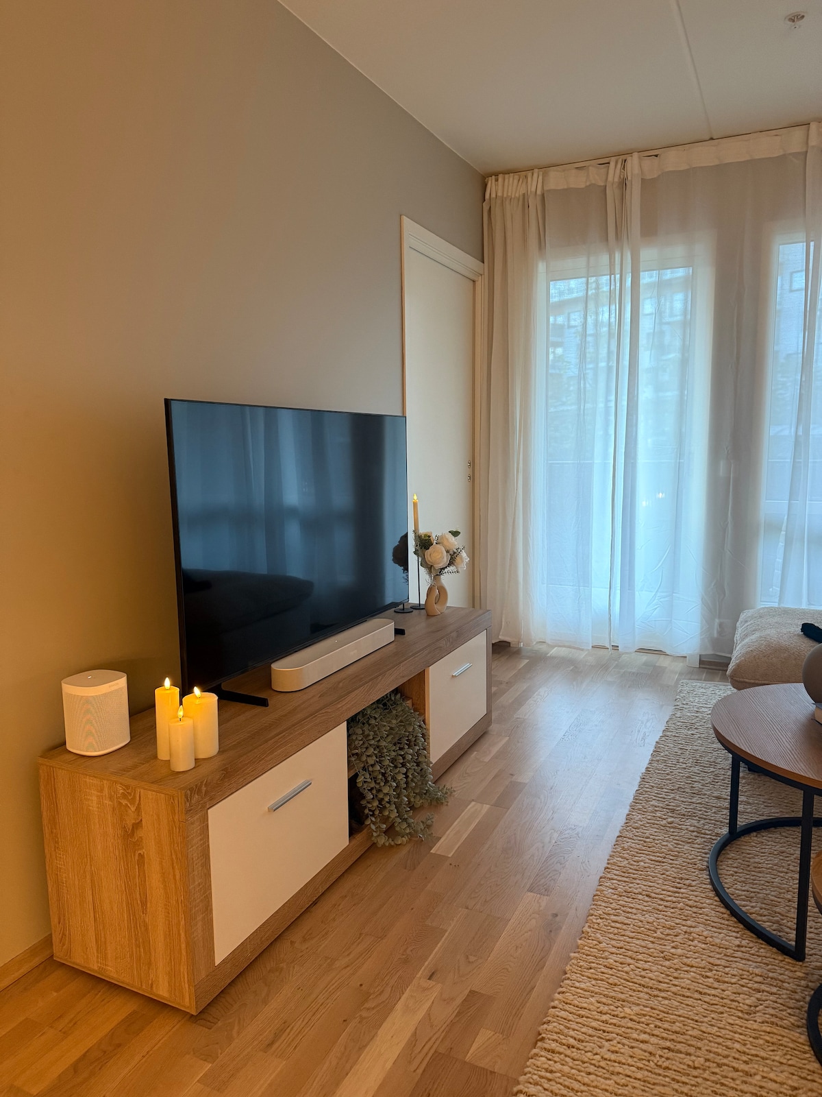 A modern living area features a large television mounted on a light wood media console. Soft lighting is provided by candles arranged on the console. Sheer curtains allow natural light to filter in, complementing the warm tones of the wooden flooring.