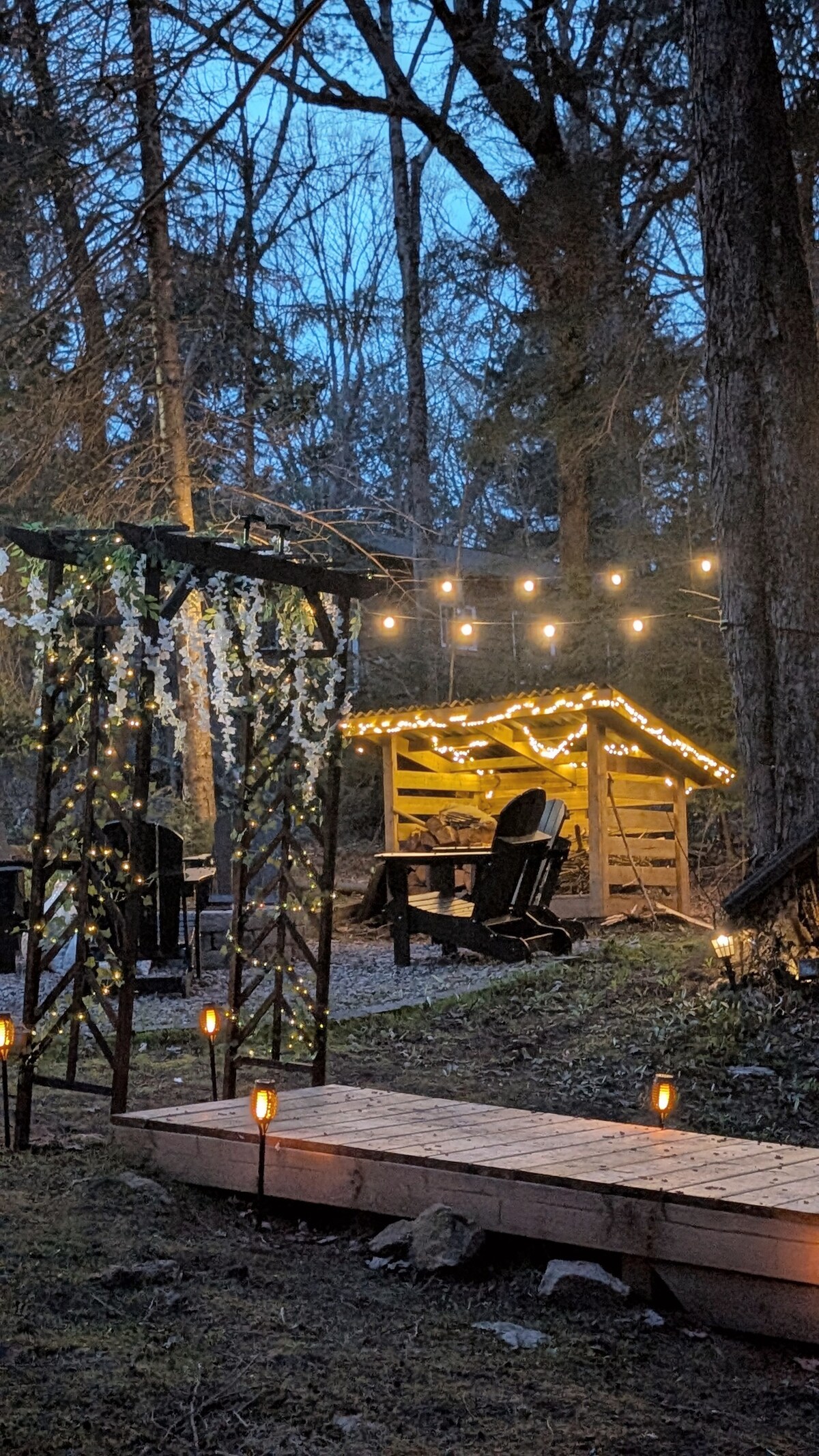 A serene outdoor space is illuminated by soft string lights, casting a warm glow among the trees. A cozy seating area features Adirondack chairs positioned near a rustic shelter. Pathways are lined with lanterns, leading through a natural setting.