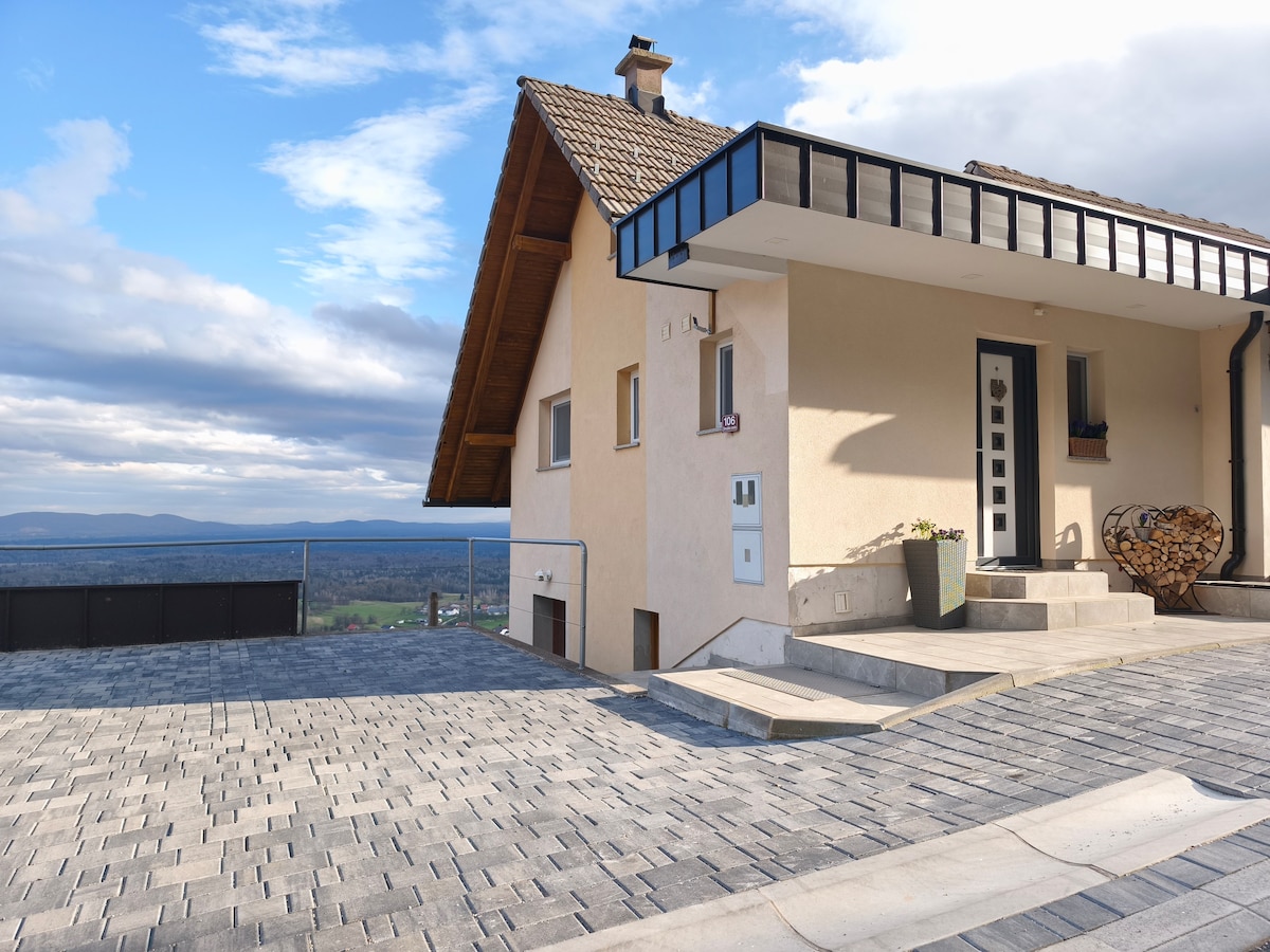 The exterior of the vacation home is depicted, showcasing a sloped roof, a front door with an inviting entryway, and a stone patio. A fenced area surrounds a cozy seating space, with wood logs neatly stacked nearby. Scenic vistas extend into the background.
