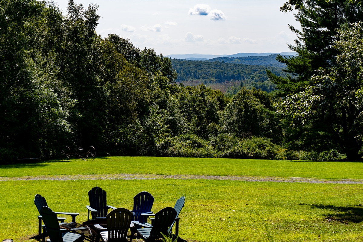 An open green space is highlighted with several Adirondack chairs arranged around a fire pit. Lush trees and vegetation surround the area, providing a serene setting. In the distance, rolling hills and mountains can be seen under a clear blue sky.