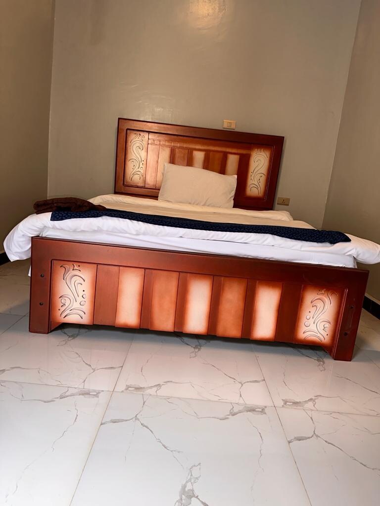 A comfortable room is featured, showcasing a wooden bed with decorative elements. Crisp white bedding is neatly arranged, and a small pillow is positioned against the headboard. The tiled floor is light-colored with subtle veining, contributing to a clean and spacious atmosphere.