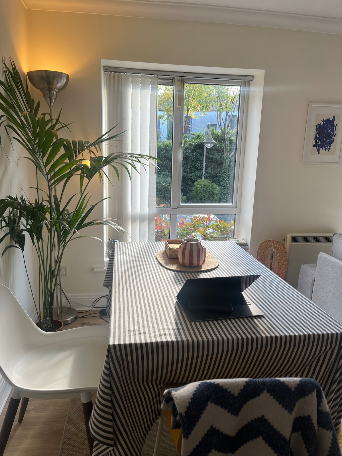 A dining area features a striped tablecloth draped over the table, accompanied by a sleek white chair. A dark tablet rests on the surface, while a decorative basket and fruit sit in the center. Natural light filters through the window, illuminating a nearby potted plant.