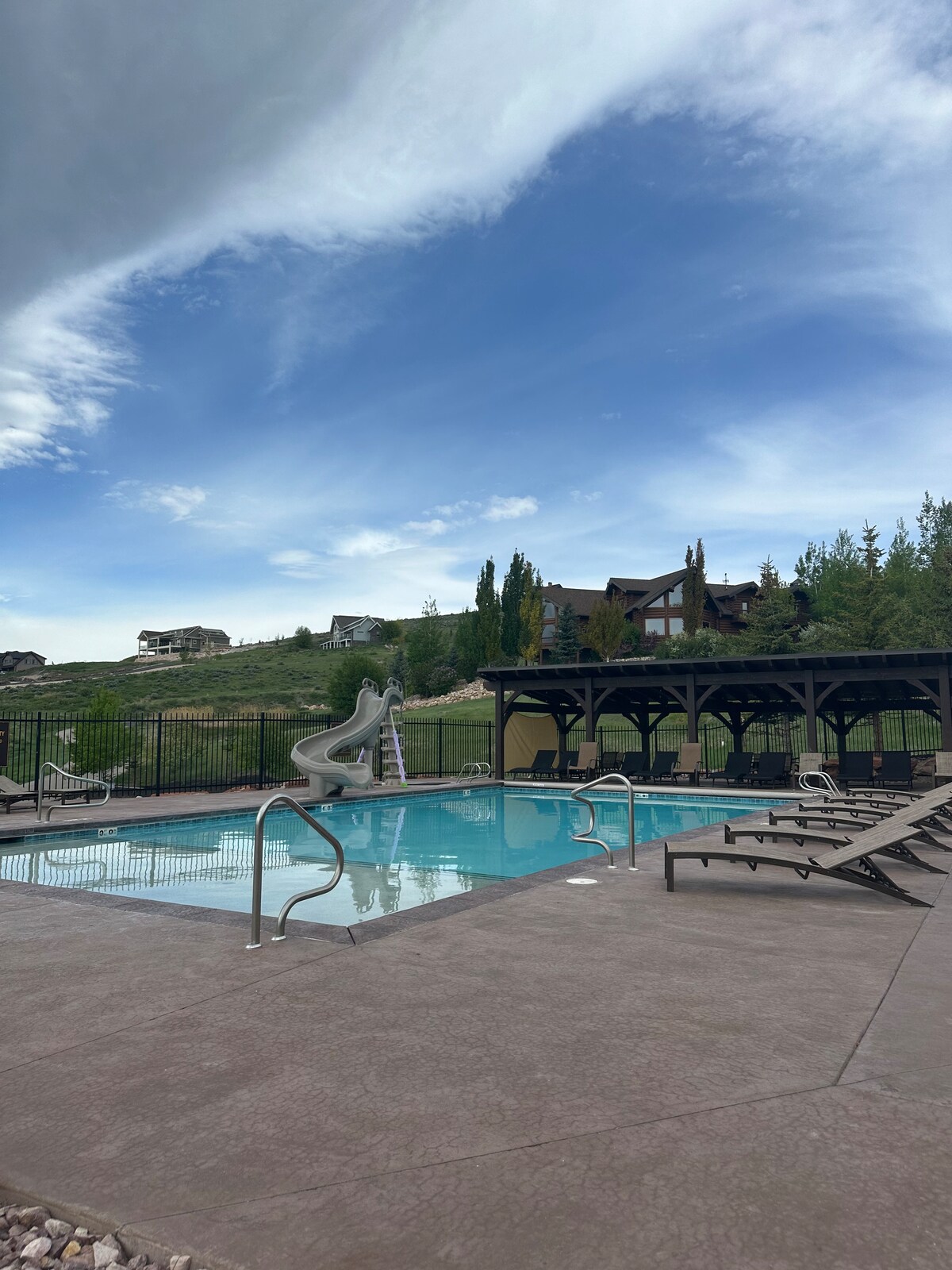A clear swimming pool is surrounded by lounge chairs, with a waterslide visible on one side. Shade is provided by a covered area, and the landscape features green hills and a distant view of nearby buildings under a clear blue sky.