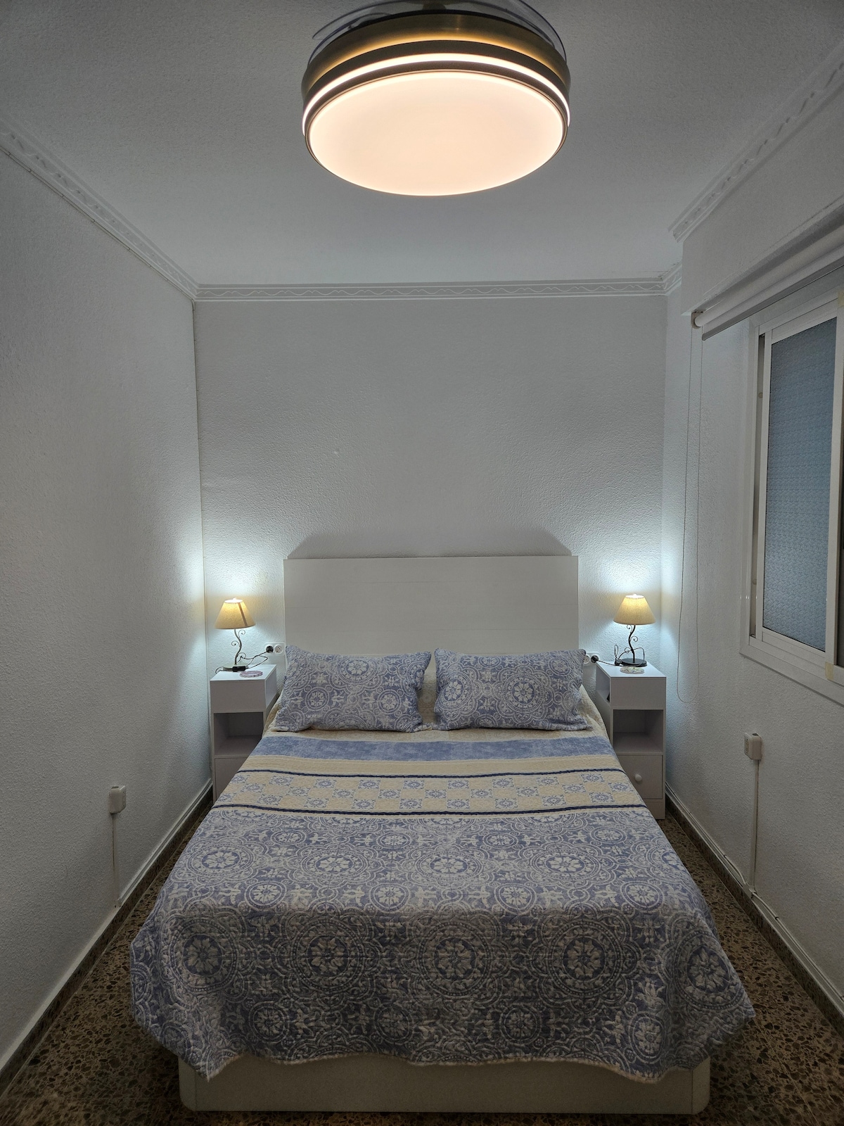 A comfortable sleeping area is presented with a spacious bed dressed in a blue patterned coverlet. Soft light is emitted from bedside lamps on either side. The room features white walls and a window that allows for natural light, enhancing the overall brightness.