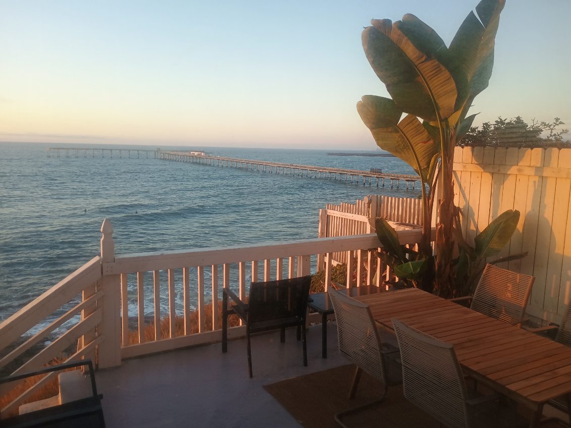 An outdoor deck offers views of the ocean and a nearby pier under the warm glow of sunset. A dining table with several chairs is positioned on the deck, along with a lounge chair. Lush palm leaves add a tropical touch to the scene.
