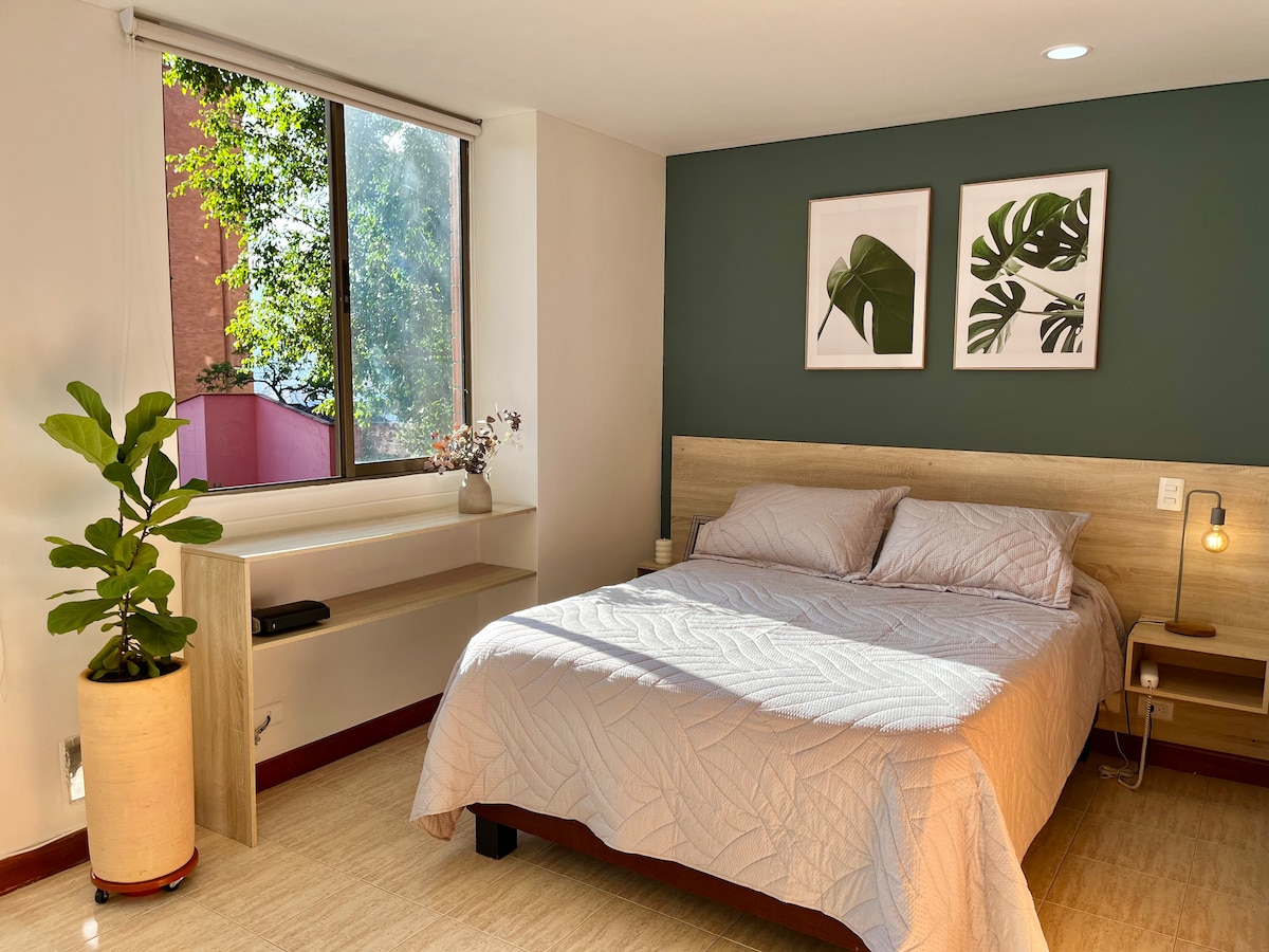 A spacious sleeping area features a comfortable bed with a light quilt. Two framed botanical prints adorn the walls. Natural light fills the room through a large window, with a small indoor plant placed beside a simple wooden shelf.