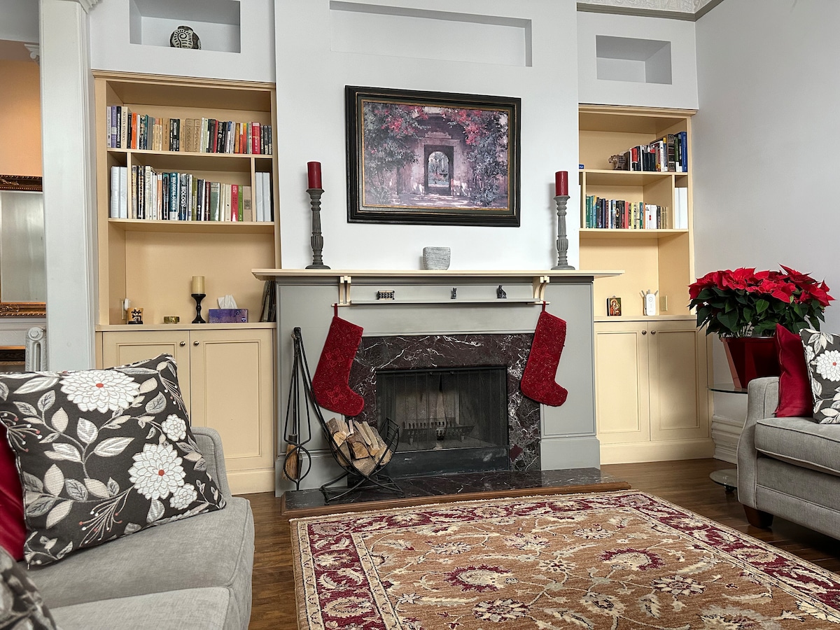 A cozy living area features a marble fireplace adorned with decorative stockings. Shelves filled with books are positioned on either side. A large framed artwork hangs above the fireplace. A patterned area rug lays on polished wooden flooring, complemented by plush seating.