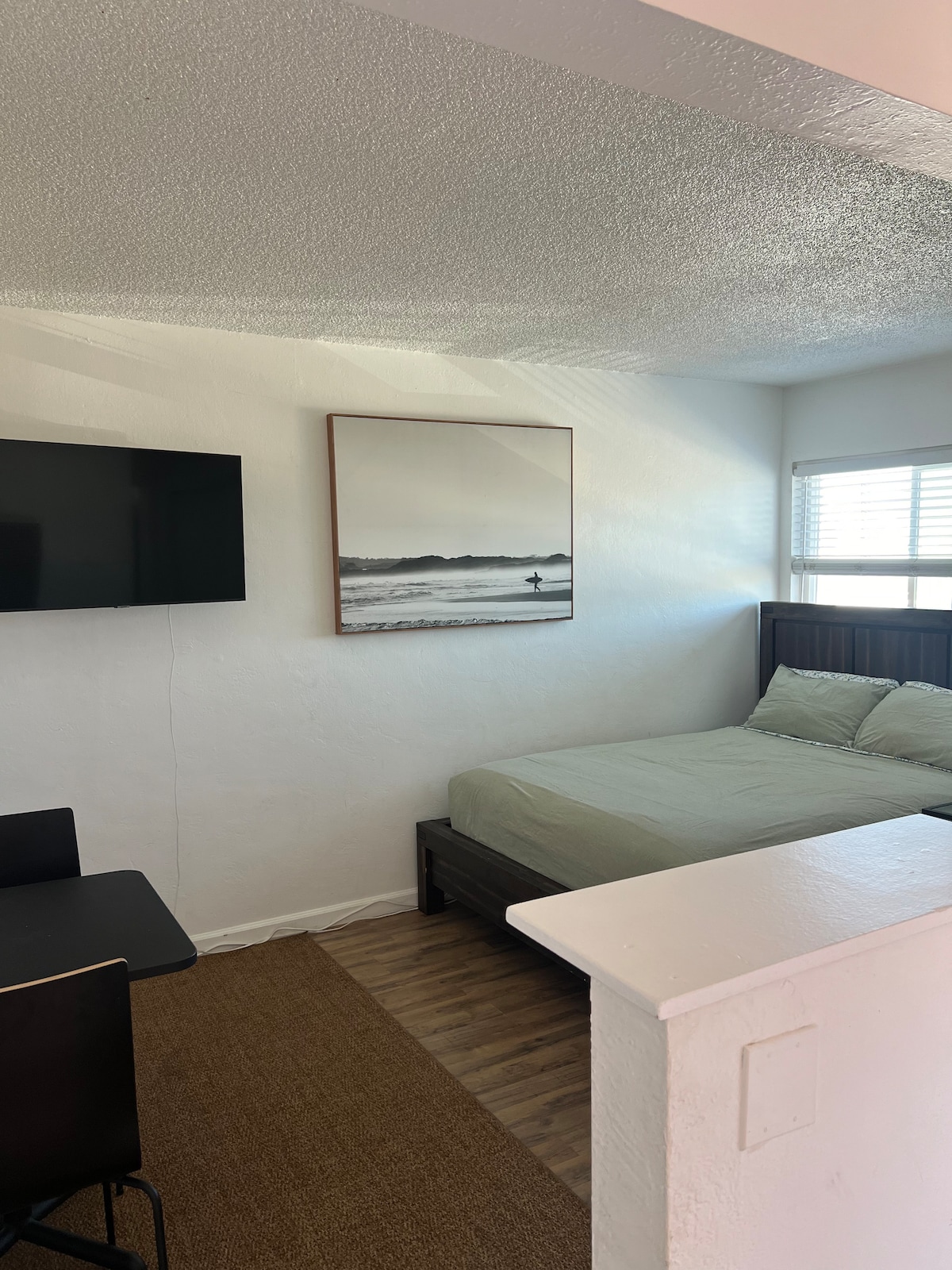 A functional studio space features a queen size bed against a wall, complemented by a large artwork depicting a serene beach scene. A flat-screen TV is mounted above a small dining table set for two, with natural light filtering through the window.