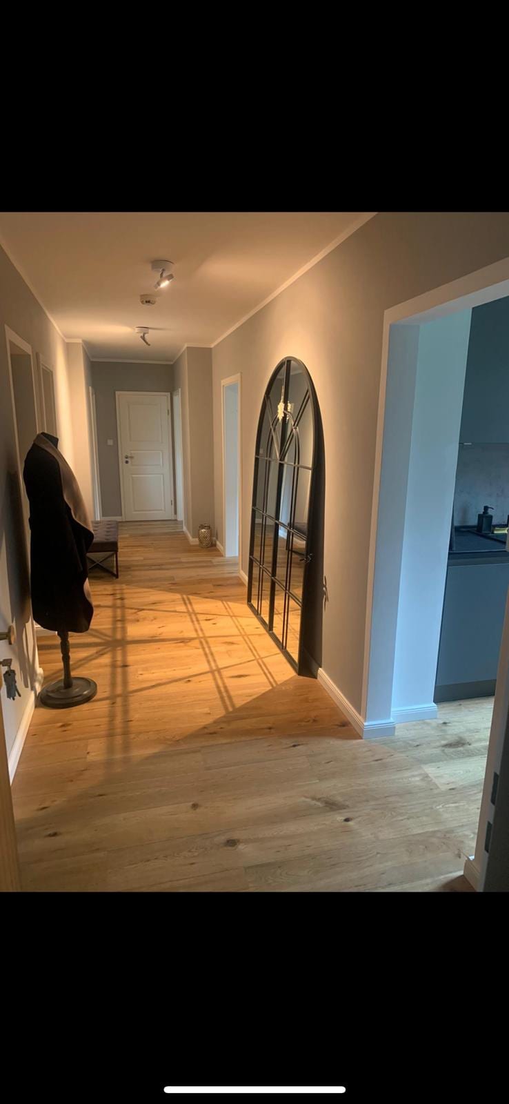 A well-lit hallway features light wood flooring and calming gray walls. An arched mirror is mounted on the wall, reflecting the space. A coat stand is positioned against the wall, while a doorway leads to a kitchen area at the end of the hallway.