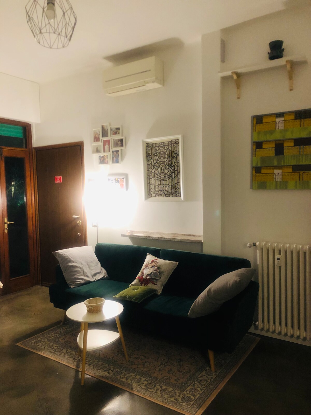A modern sitting area features a plush green sofa with decorative pillows and a small side table. Soft lighting from a standing lamp enhances the cozy ambiance. A colorful wall art piece and framed photographs adorn the walls, adding character to the space.