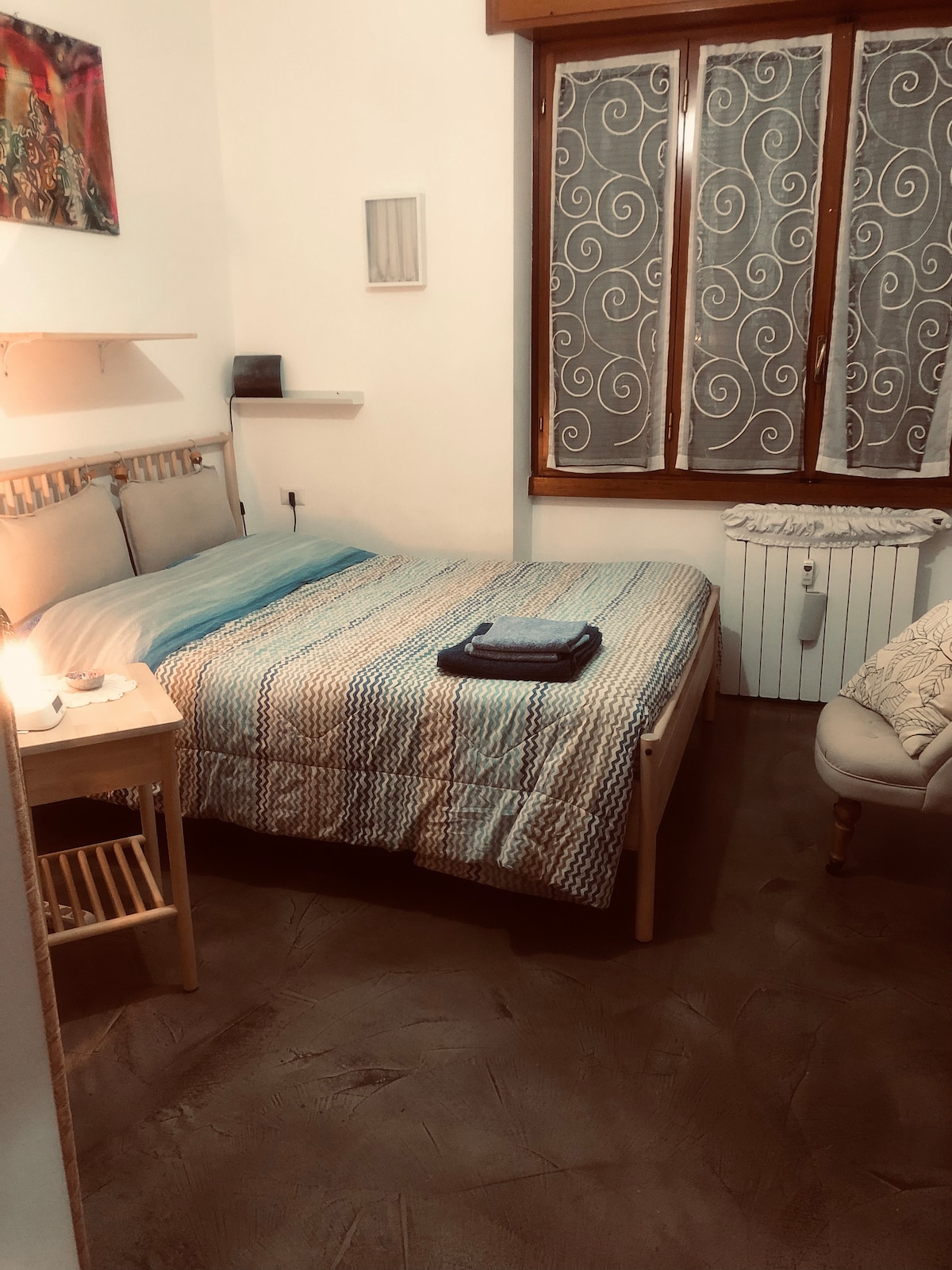 A neatly arranged bedroom features a spacious bed dressed in a textured quilt with blue and beige tones. A small bedside table holds a lamp, while a cozy armchair is positioned nearby. Natural light enters through a curved window with patterned curtains, enhancing the calming atmosphere.