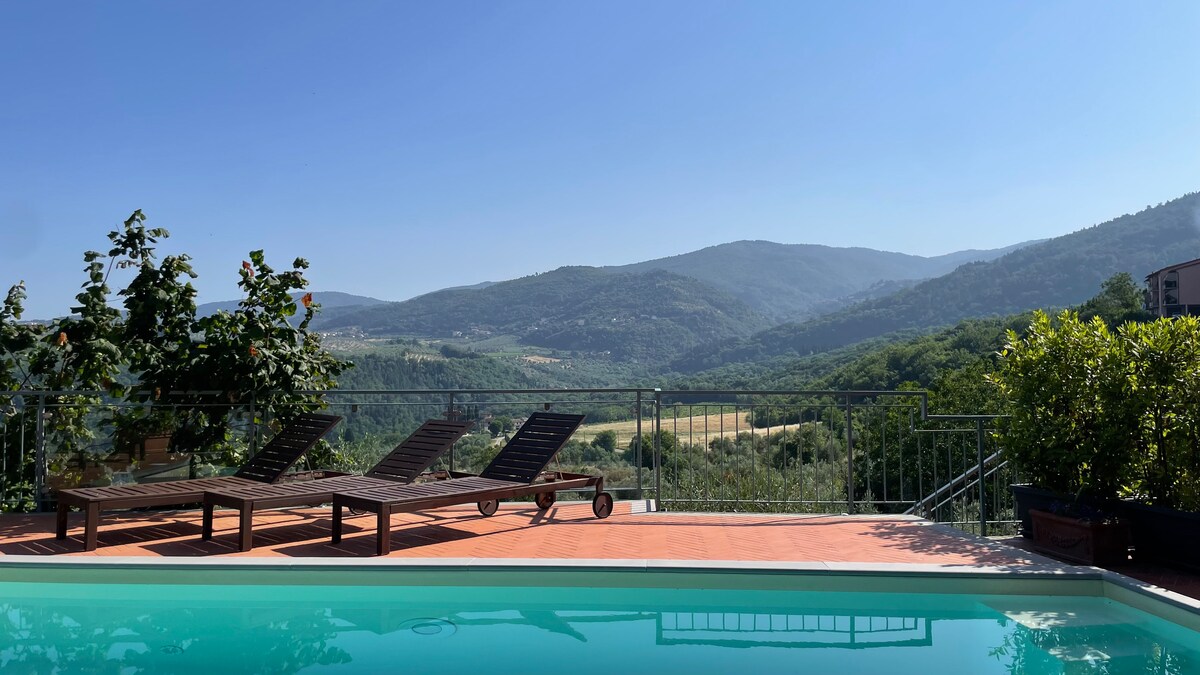 A serene outdoor area features a private pool surrounded by sun loungers. A clear view of rolling hills and greenery extends into the distance under a bright blue sky, showcasing the peaceful nature of the landscape.