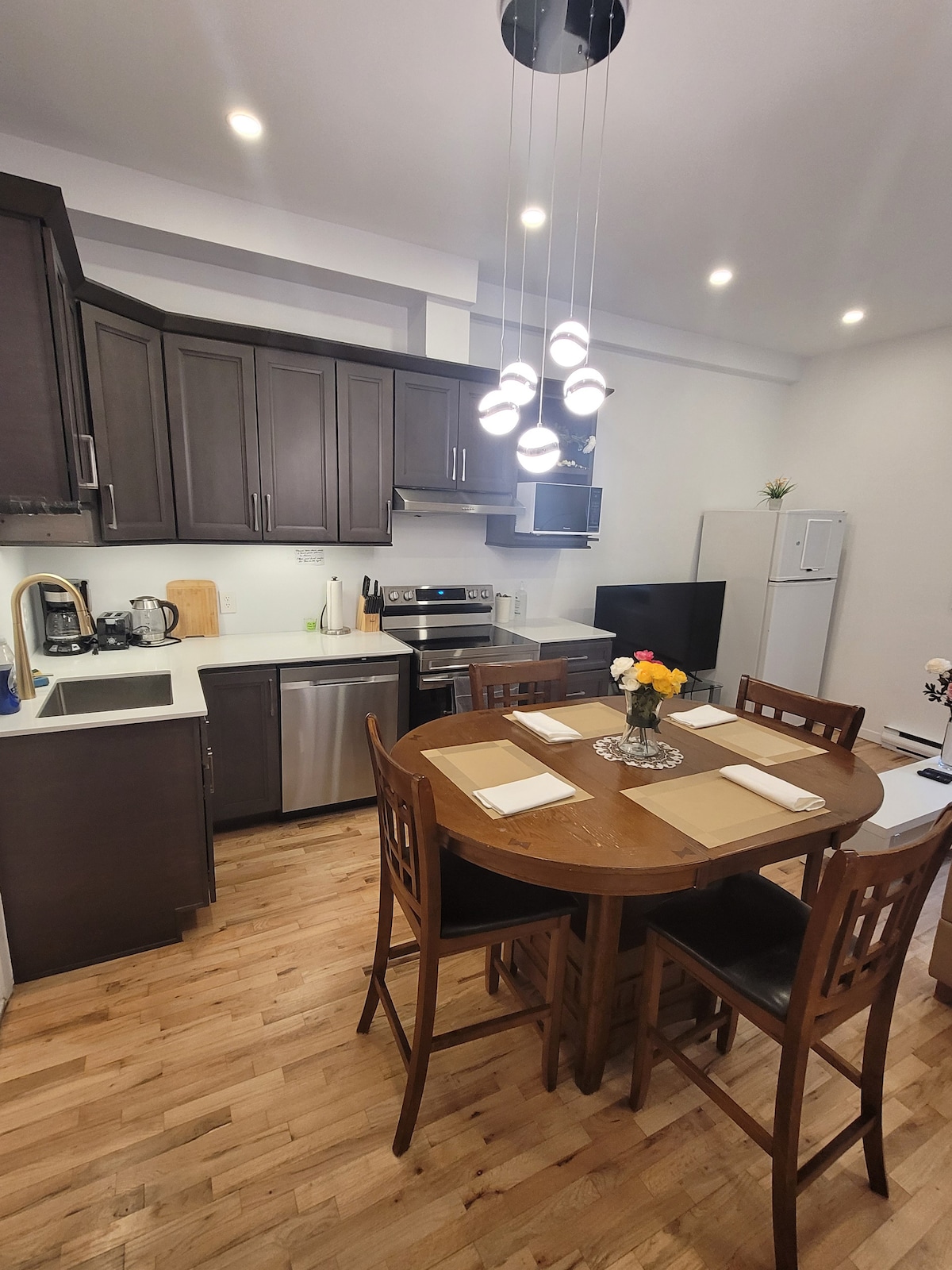 The kitchen features modern cabinetry and stainless steel appliances, including a stove and refrigerator. A circular dining table with four chairs is centrally positioned, adorned with a small vase of flowers. Soft lighting enhances the warm atmosphere throughout the space.