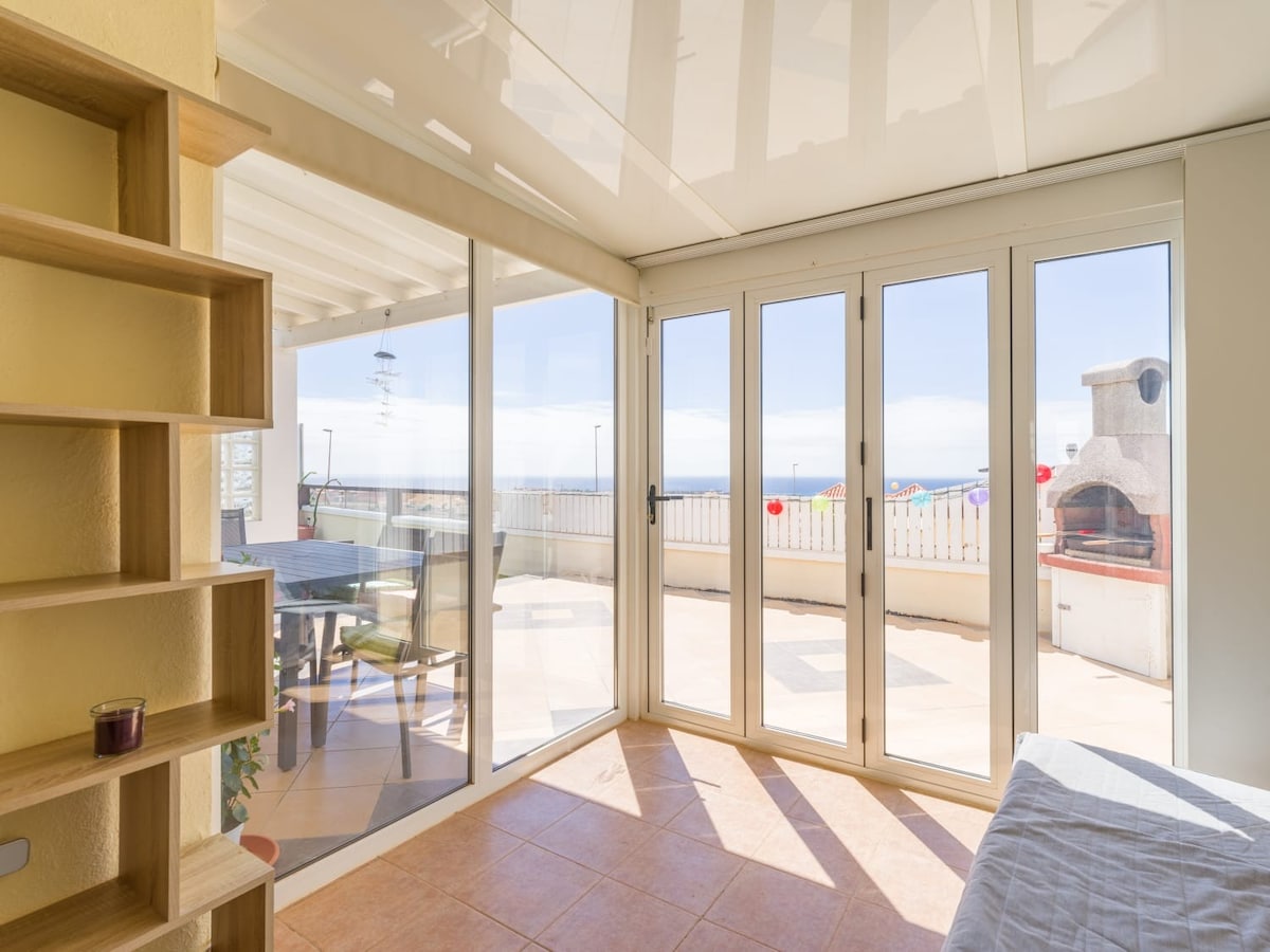 A bright interior space is framed by large glass doors, offering views of a spacious terrace and the ocean. Sunlight fills the room, highlighting a cozy seating area and a bookshelf. A distinct outdoor grilling area is visible in the background.