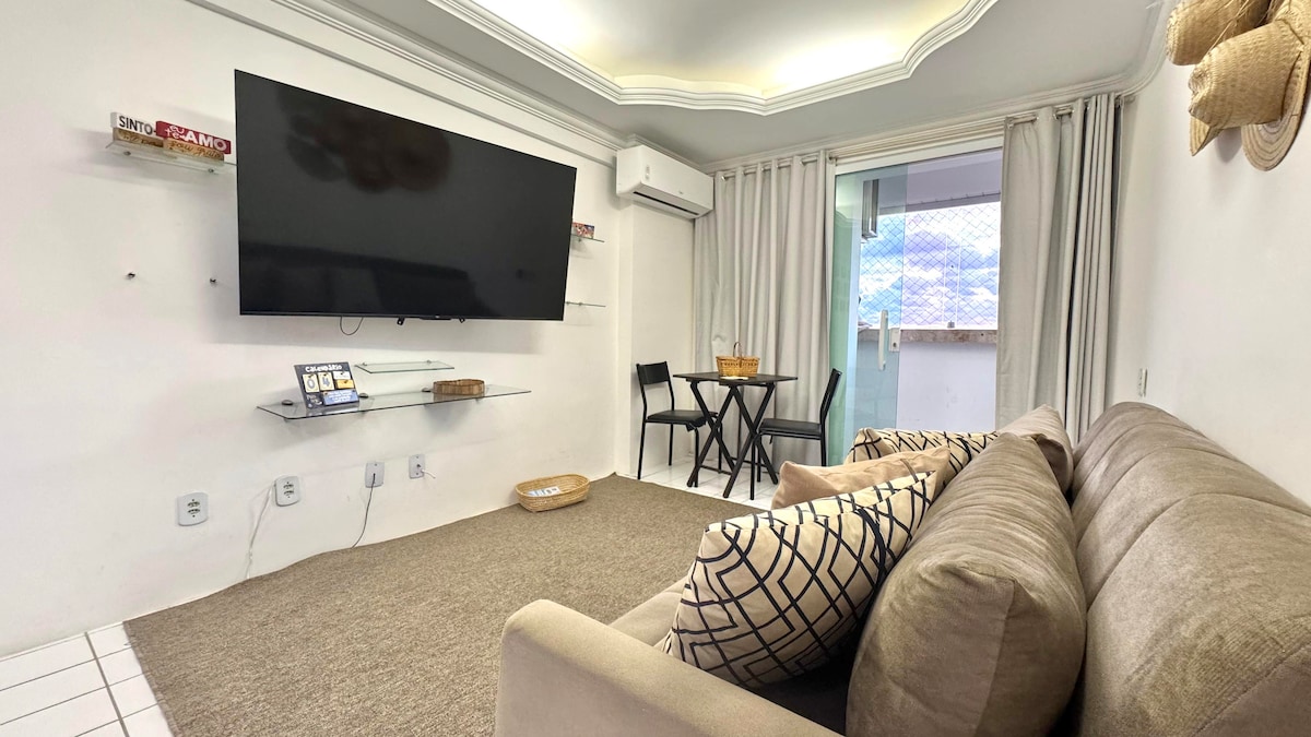 A cozy living area features a comfortable sofa and an entertainment system mounted on the wall. A small dining table with chairs is positioned nearby, set against large windows that allow natural light to fill the space.