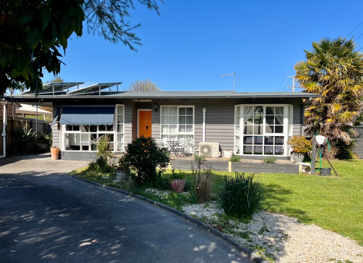 The cottage exterior features a grey façade with white trim, showcasing large windows and a welcoming orange front door. Lush green landscaping surrounds the property, including a mixture of plants and flowers, while a well-maintained lawn extends towards the driveway.