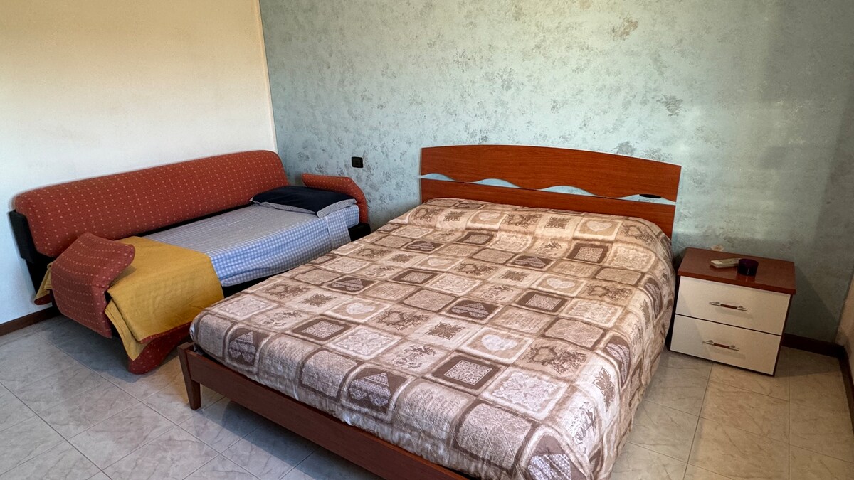 A inviting bedroom features a spacious bed with a textured coverlet. A sofa bed is positioned beside it, offering additional seating and sleeping options. A bedside table holds a small lamp, complementing the light-colored floor and softly colored walls.