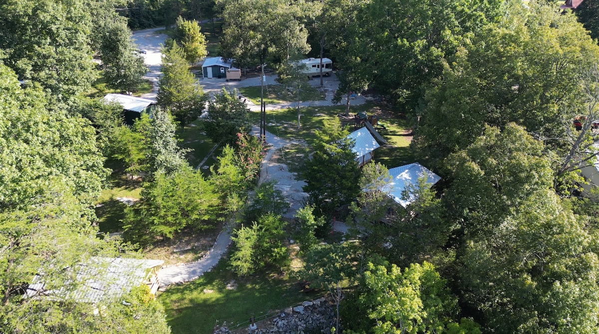 An aerial view of the glamp-ground reveals lush green trees surrounding three glamping tents and a tiny home. Gravel pathways connect the accommodations, while shaded areas invite outdoor relaxation. A well-maintained landscape blends with natural elements, creating a serene and inviting setting.