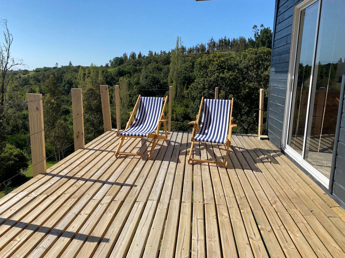 A wooden deck is shown, featuring two striped lounge chairs facing a natural landscape. The surrounding greenery is visible, providing a serene backdrop. Large glass doors lead into the interior space, allowing for ample natural light.