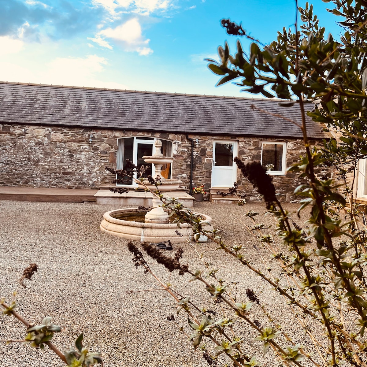 The exterior of a stone cottage is visible, featuring a simple entrance with a white door and windows. A circular stone fountain is positioned in the foreground, surrounded by gravel. Green foliage frames the scene, complementing the blue sky above.