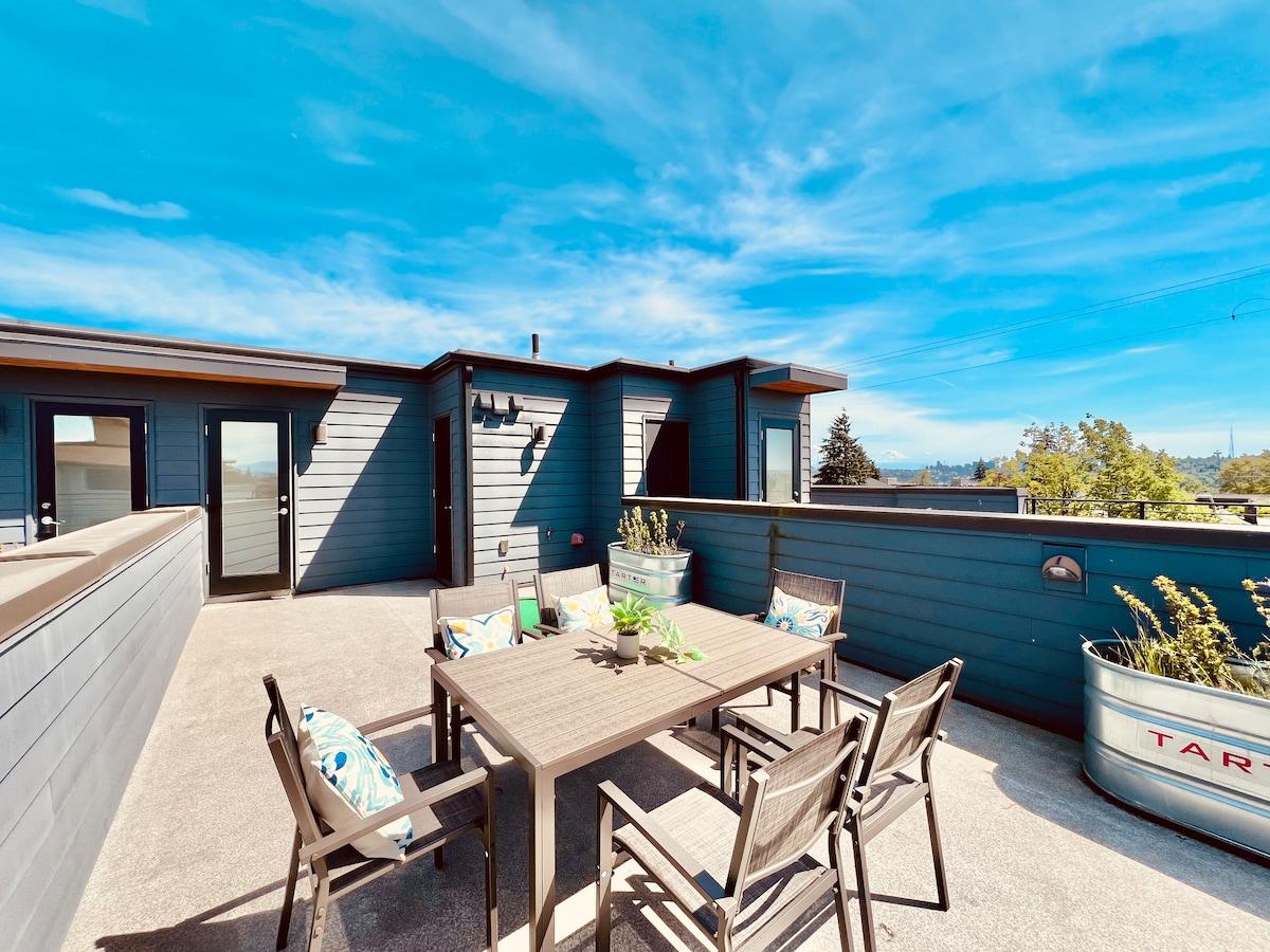 The rooftop deck is furnished with a dining table surrounded by six chairs, offering ample seating for outdoor meals. The space is surrounded by railing, providing safety. Views of the surrounding area and blue sky contribute to an inviting ambiance for relaxation.