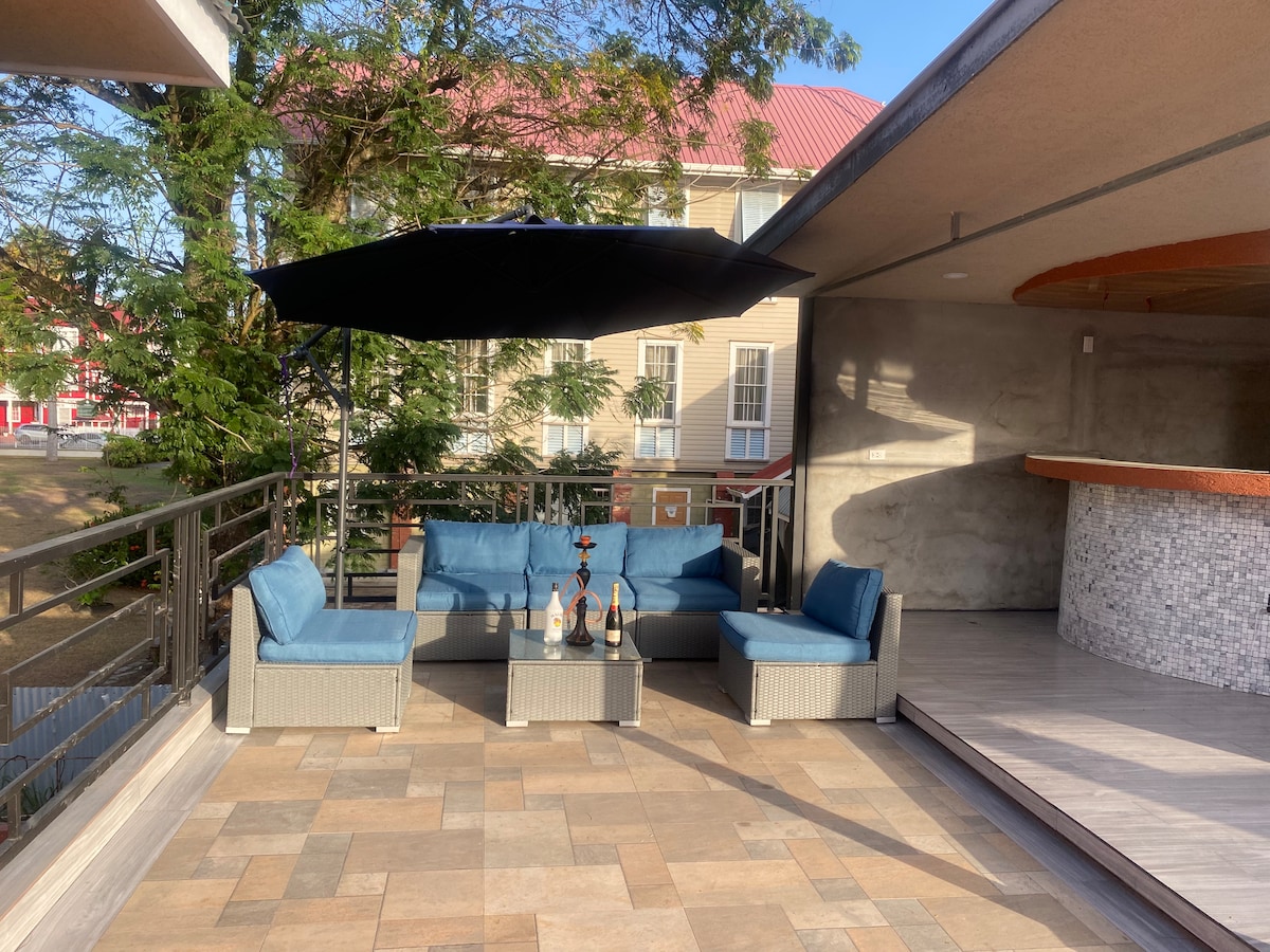 A shaded outdoor seating area is featured, furnished with a cozy arrangement of blue cushions on a textured gray sofa set. A patio umbrella provides shade, while a bar counter is visible in the background, creating an inviting space for relaxation.