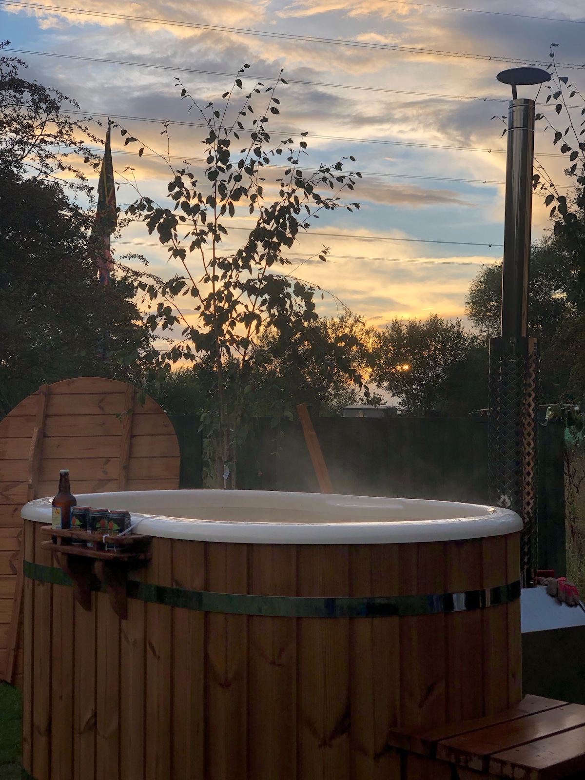 A wood-burning hot tub is set against a backdrop of a colorful sunset. Steam rises from the water, creating a serene atmosphere. Surrounding trees and a wooden privacy screen enhance the sense of seclusion, while a drink rests on the edge, inviting relaxation.