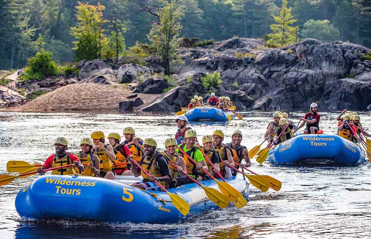 A group of colorful rafts is visible on a calm river, surrounded by rocky shorelines and lush greenery. Participants in life jackets are seated in the rafts, engaged in the outdoor activity. The sky is clear, enhancing the natural surroundings.